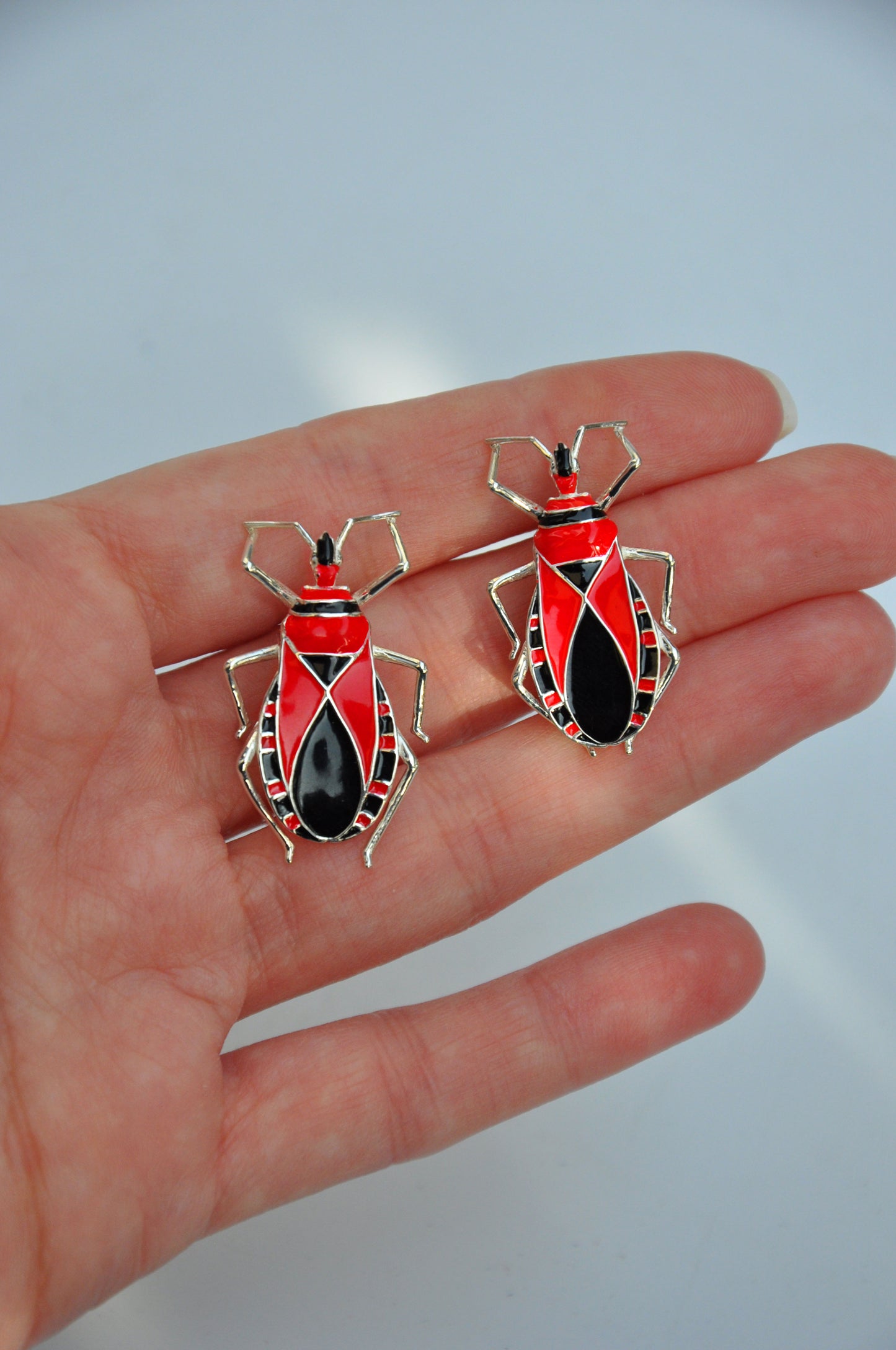 Sterling silver Assassin Bug stud earrings with red and black enamel, shown held in hand to highlight their sculptural insect‑inspired design.