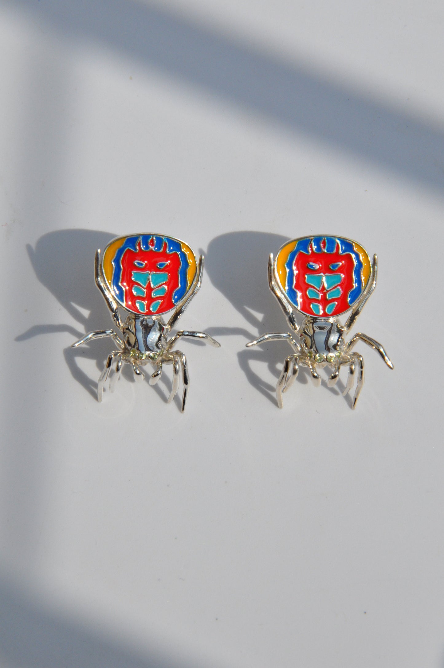 Pair of peacock spider earrings with vibrant enamel detailing displayed on a white background.