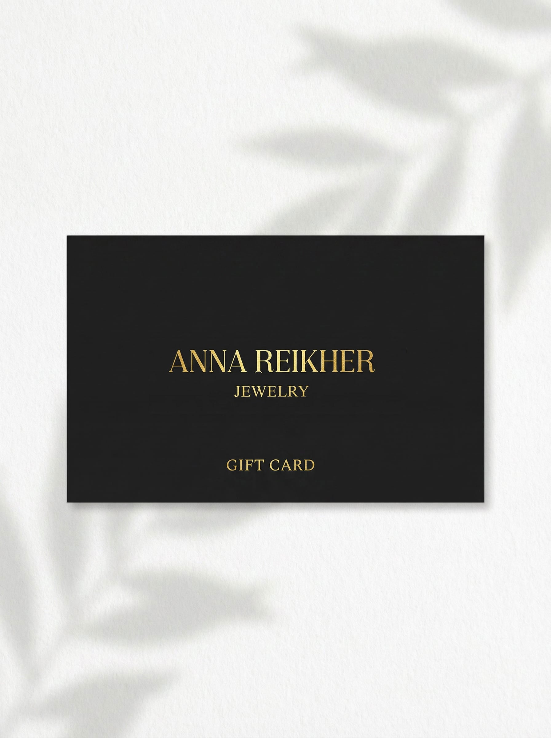 Black and gold Anna Reikher Jewelry gift card photographed on a light surface with soft leaf shadows.
