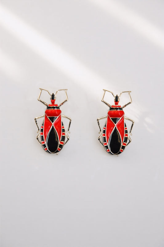 Pair of Assassin Bug stud earrings with red and black enamel detailing, crafted in metal with sculptural insect‑inspired design.