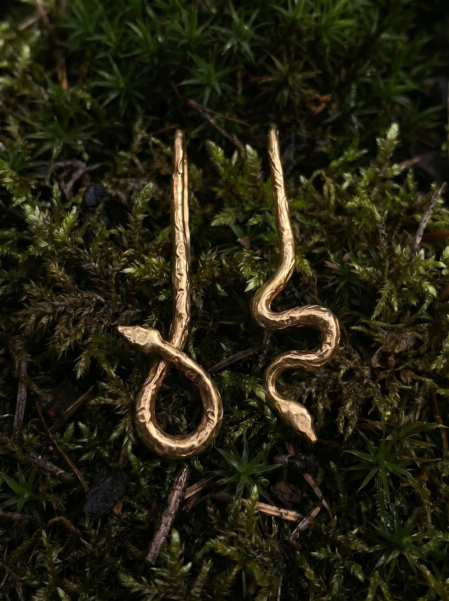 Pair of handcrafted gold serpent earrings with engraved texture, displayed on natural moss background.