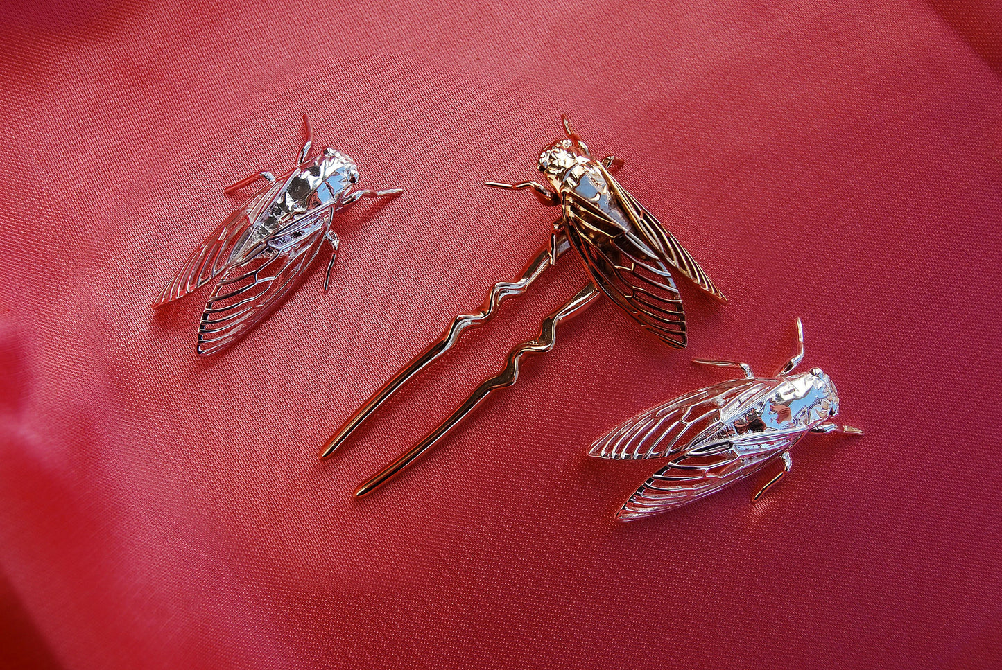 Bronze ant-shaped hair pin and silver cicada pendants arranged on soft silk fabric, highlighting the intricate insect designs and contrasting metallic textures against the smooth, luxurious backdrop.