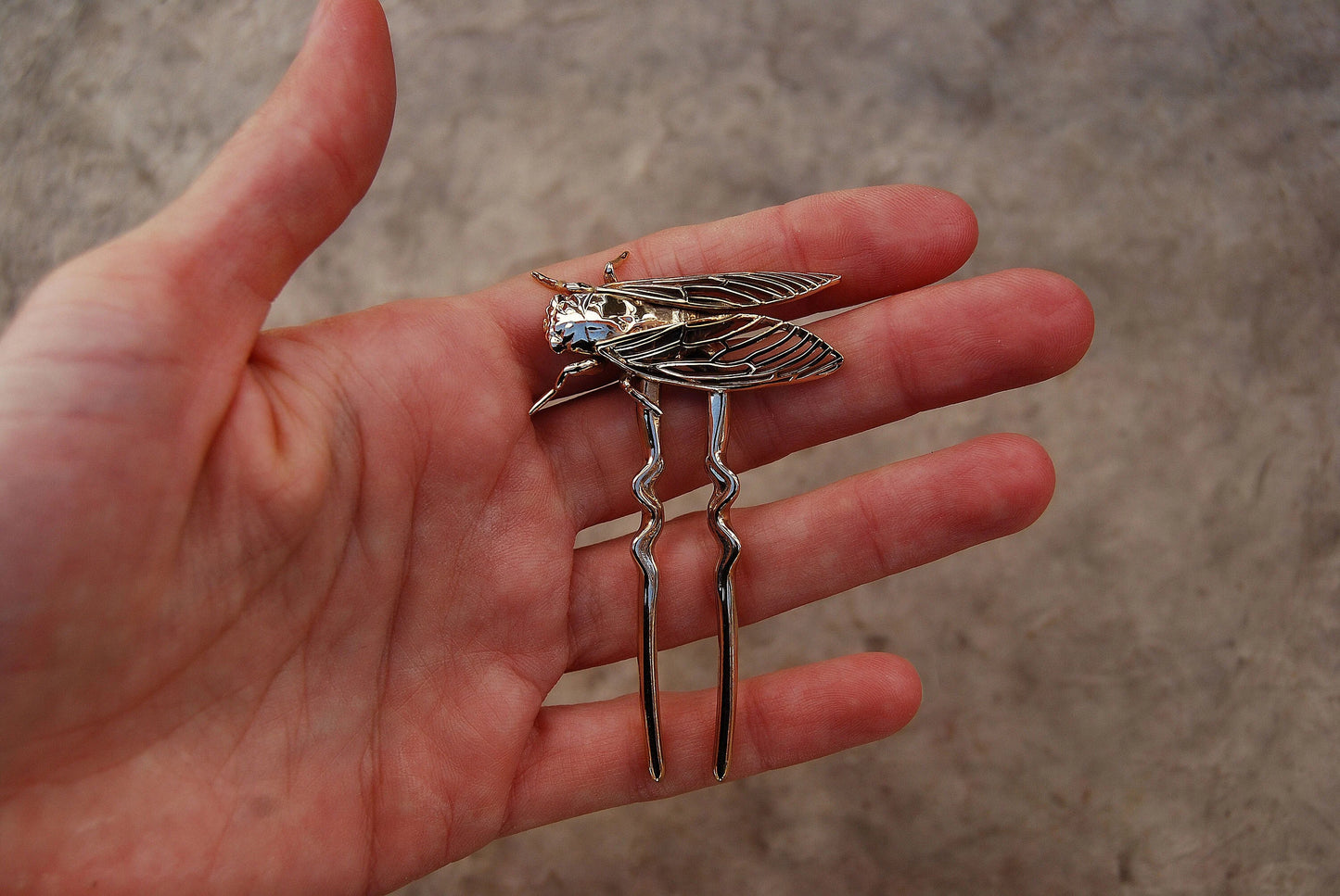 Close-up of a hand delicately holding a bronze cicada hair pin, showcasing the detailed insect design and metallic finish against a softly blurred background.