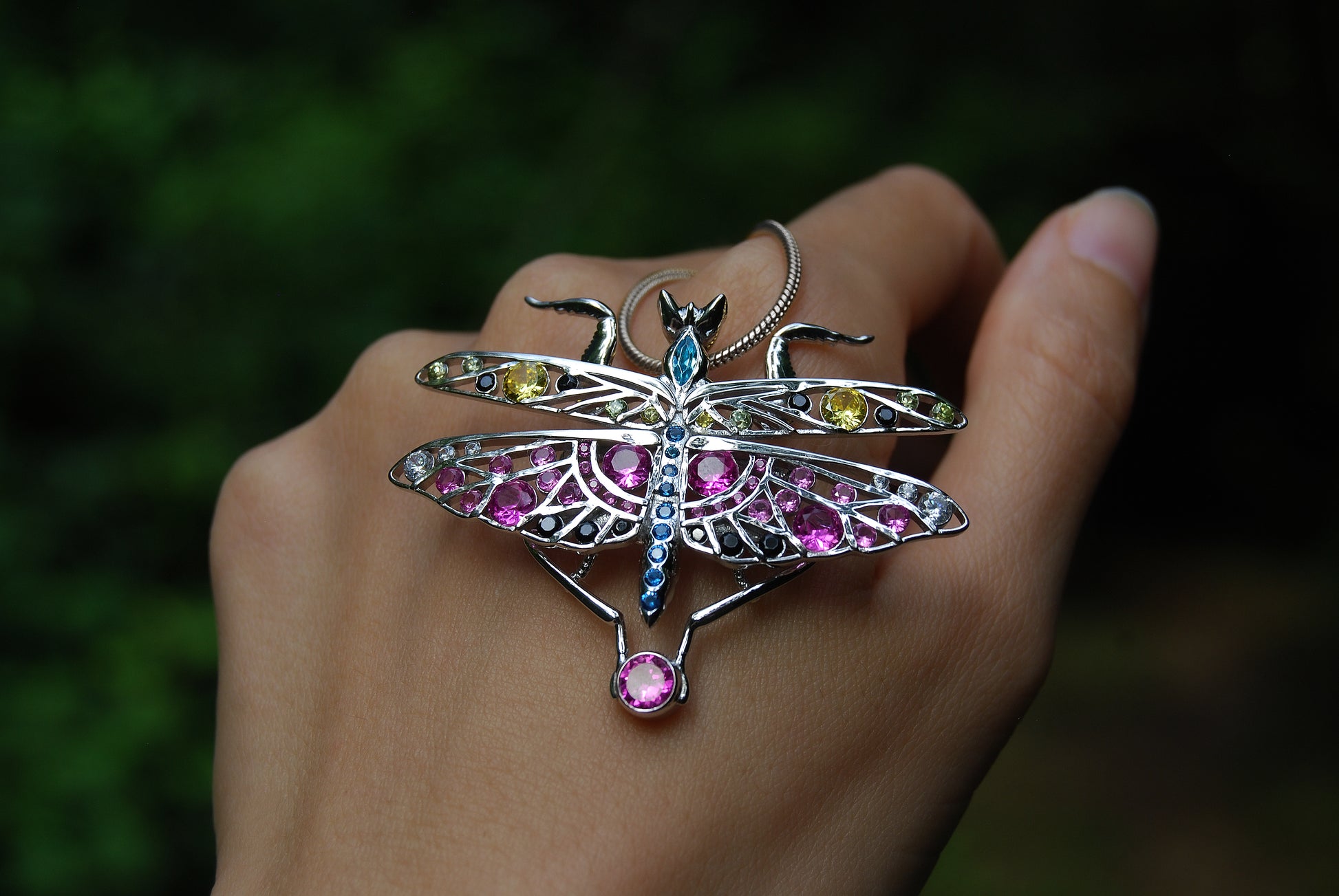 Close-up of silver praying mantis jewelry resting on hand in natural outdoor setting with soft sunlight and earthy background