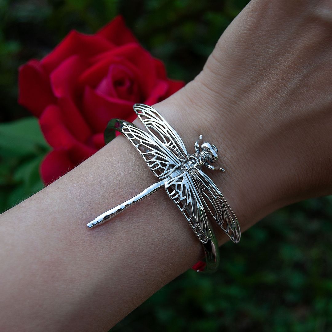 A bracelet featuring a highly detailed silver dragonfly with a silver plating, displayed on a person's wrist next to a red rose.