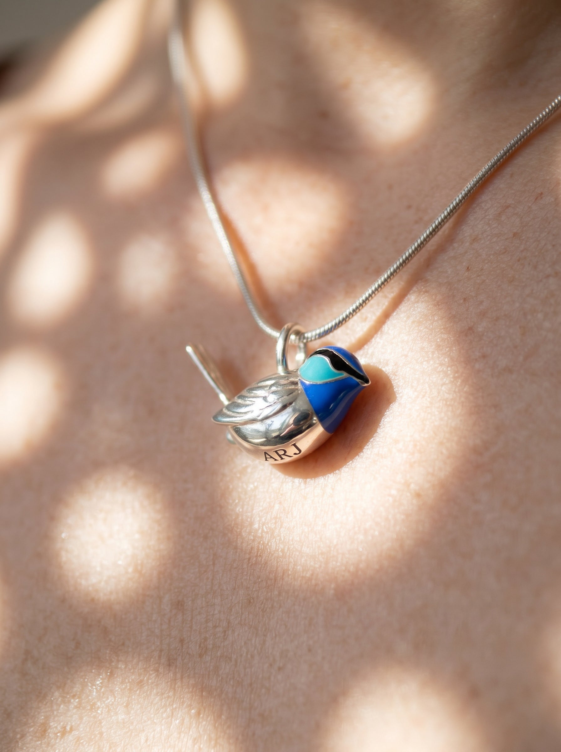 Close-up of Fairy Wren pendant worn on neck, showing sculpted silver bird with blue enamel head and engraved ARJ detail on the body.