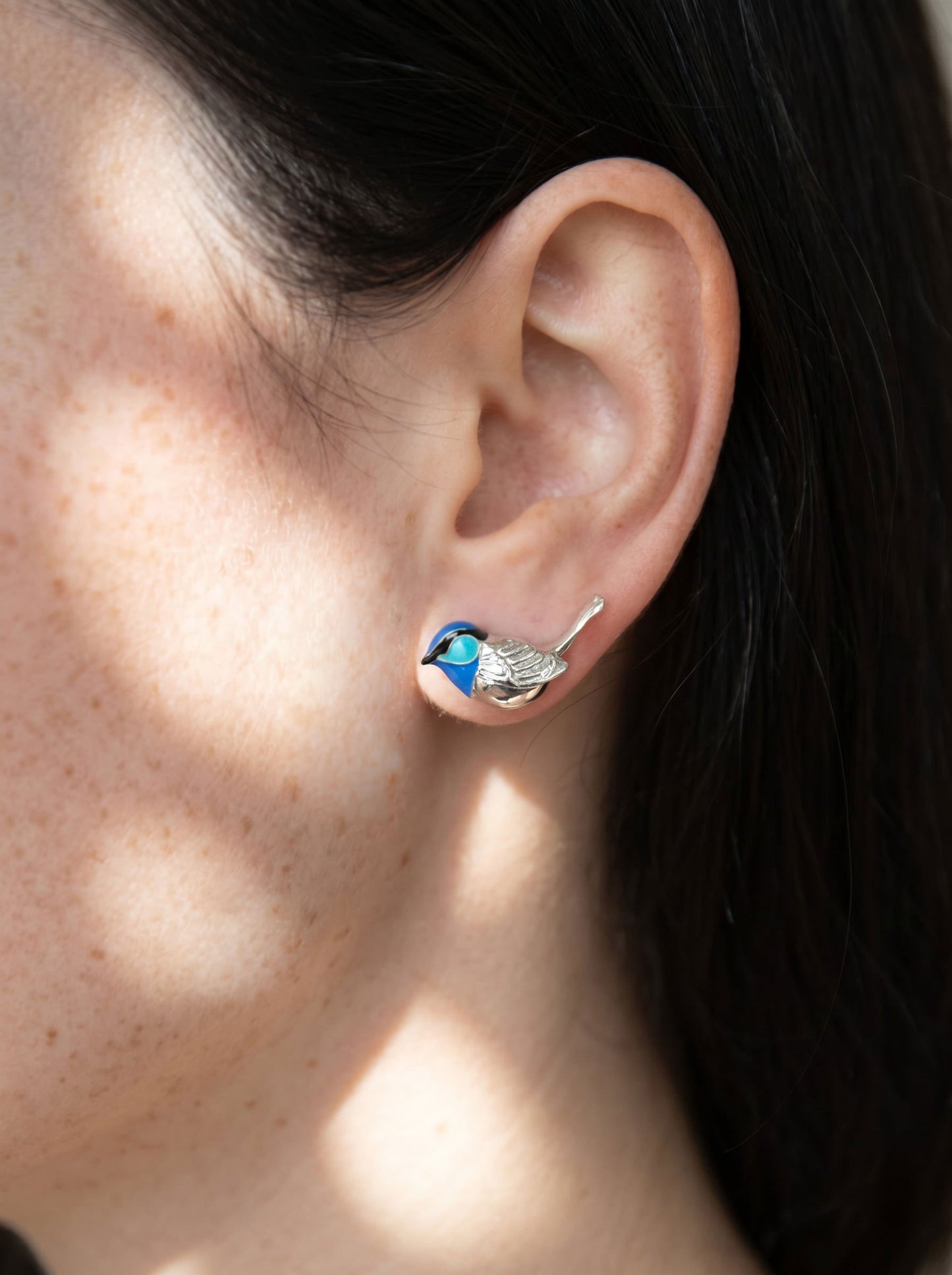 Close-up of Fairy Wren stud earring worn on ear, showing sculpted silver bird with vivid blue enamel head and fine tail detail.