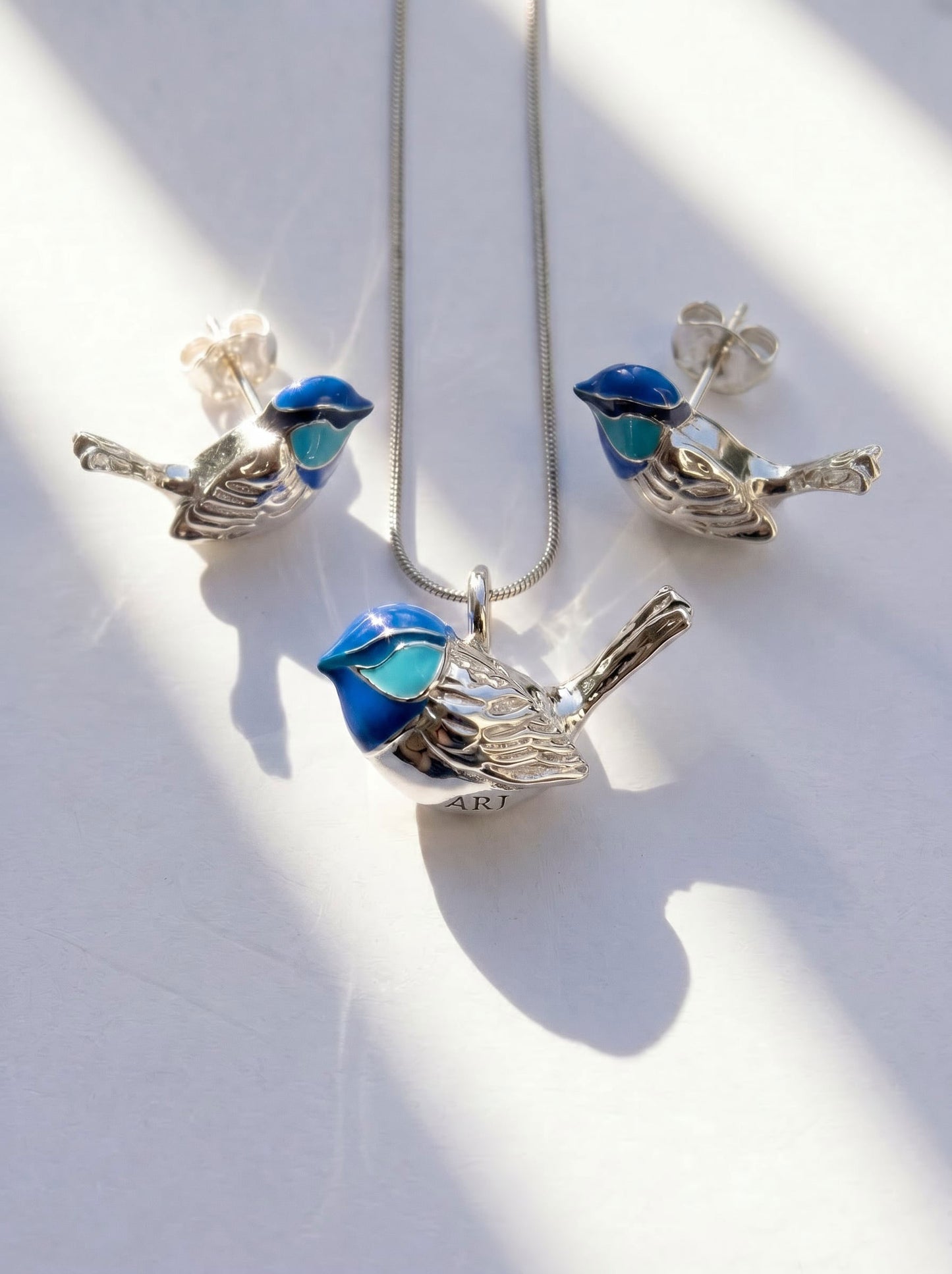 Fairy Wren stud earrings and matching necklace arranged on white surface, with sunlight casting gentle shadows across silver and enamel.