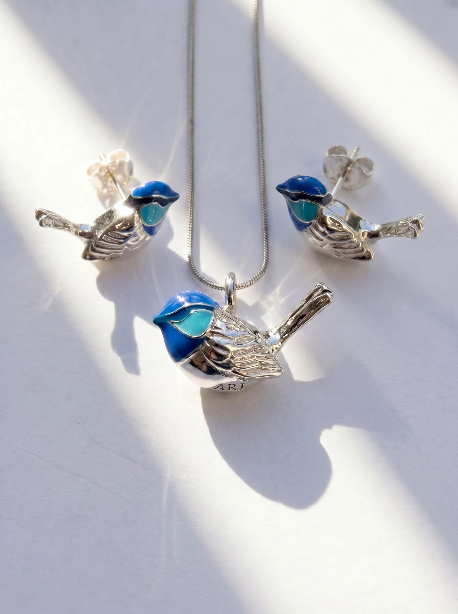 Fairy Wren stud earrings and matching necklace arranged on white surface, with sunlight casting gentle shadows across silver and enamel.