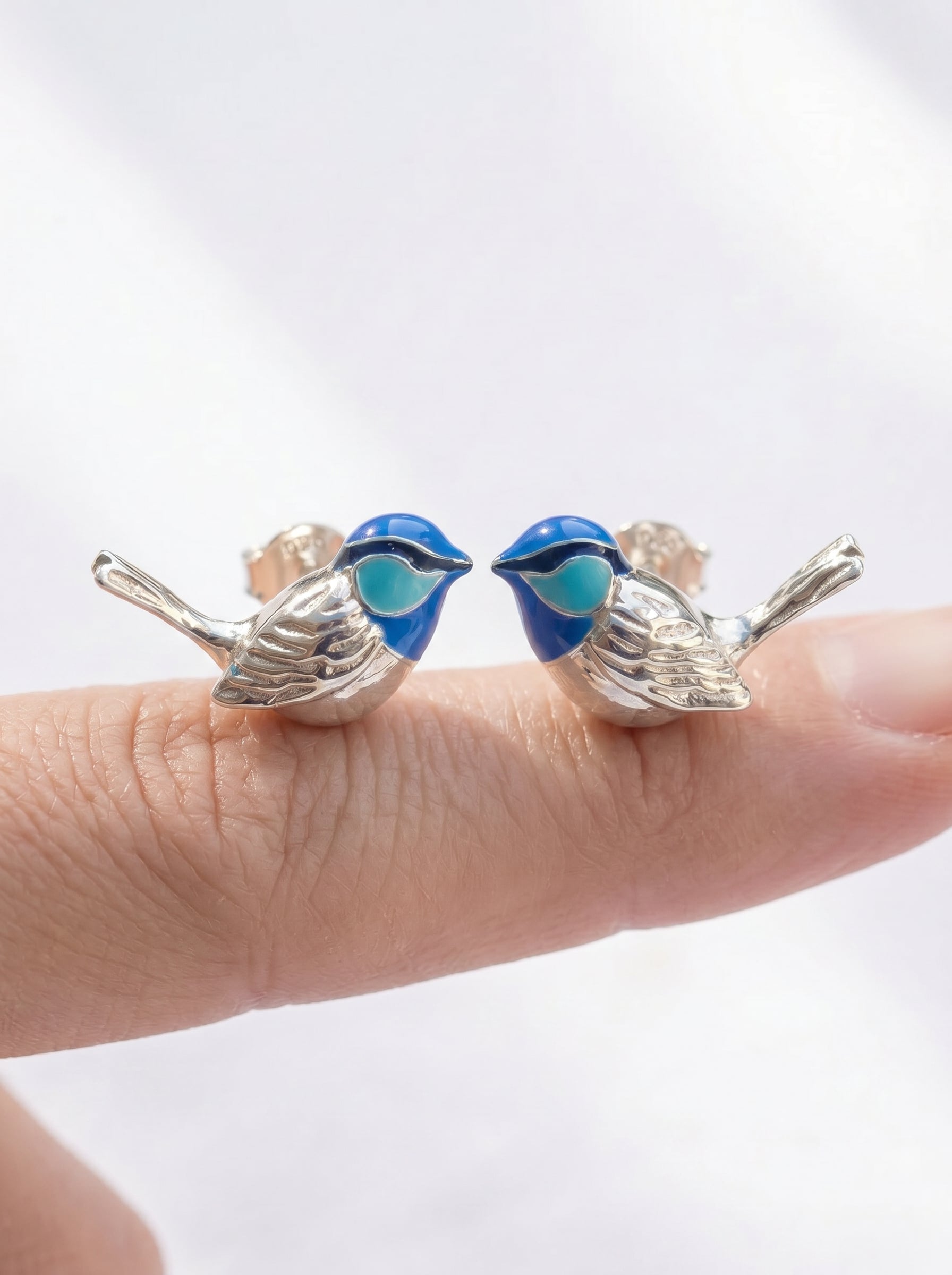 Close‑up of Fairy Wren stud earrings held between fingers, showing fine tail engraving and luminous blue enamel.