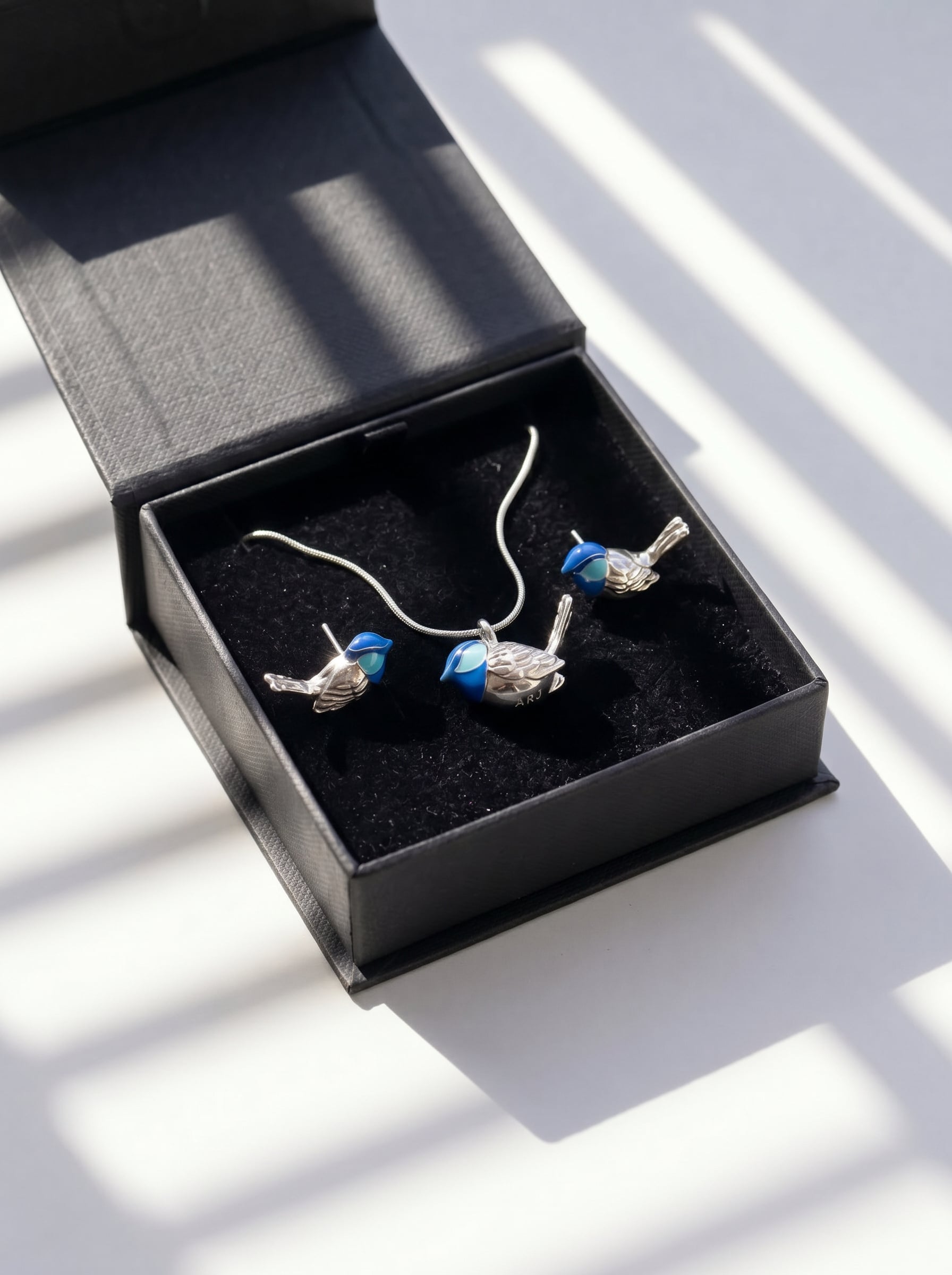 Fairy Wren pendant displayed in black jewelry box, with sunlight casting soft shadows across silver and enamel surfaces.