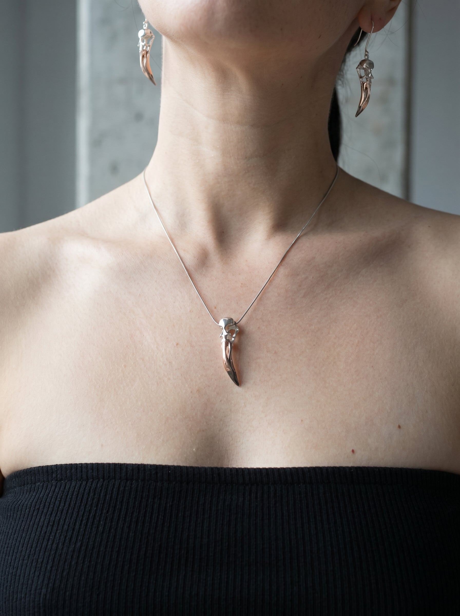 Close-up of Flamingo Skull Drop Earrings and matching pendant worn together, showing elongated sculptural skull forms in sterling silver with black rhodium and rose-gold accents against a black top.