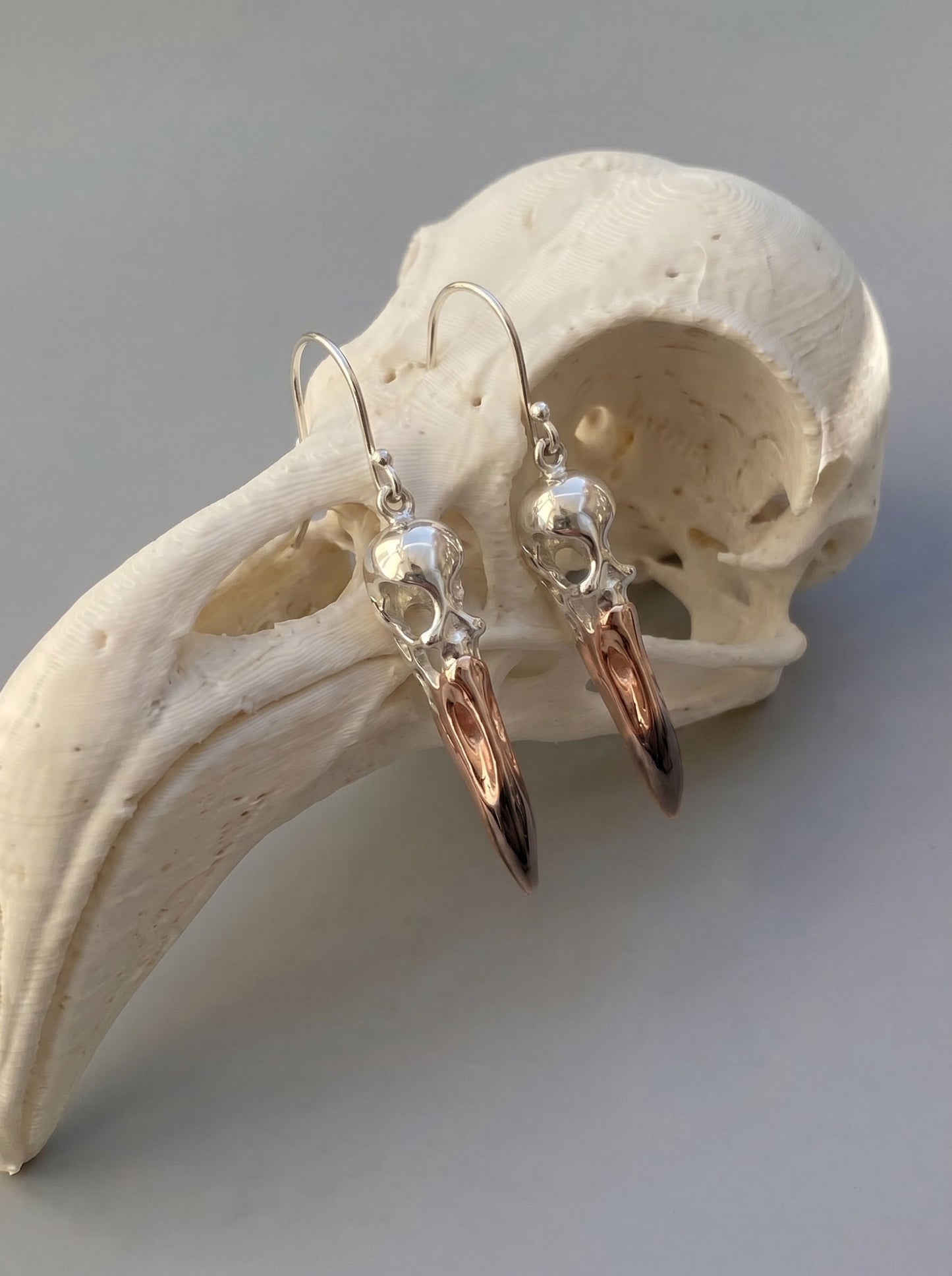 Flamingo Skull Drop Earrings displayed on sculptural stand, showing elongated beak design and warm metallic reflections.