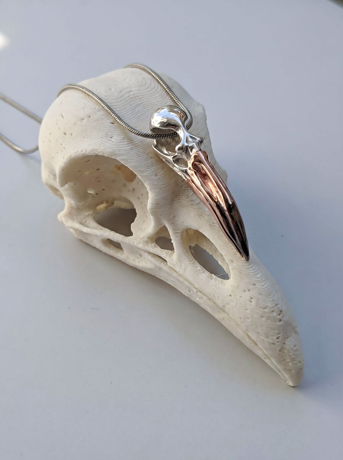 Flamingo Skull Pendant in sterling silver with black rhodium and rose‑gold accents, displayed on sculptural bird skull stand under soft light.