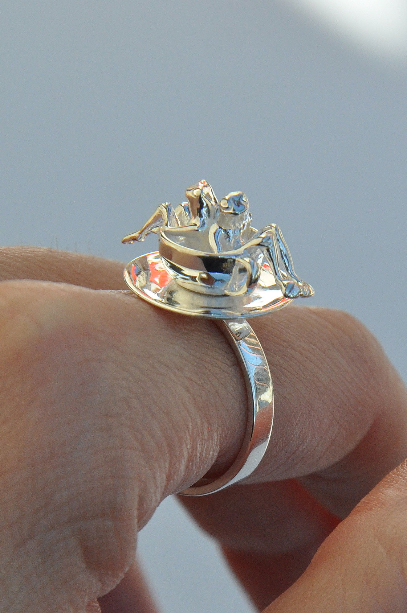 Whimsical kinetic ring worn on a finger, featuring two tiny frogs lounging inside a silver teacup atop a spinning saucer