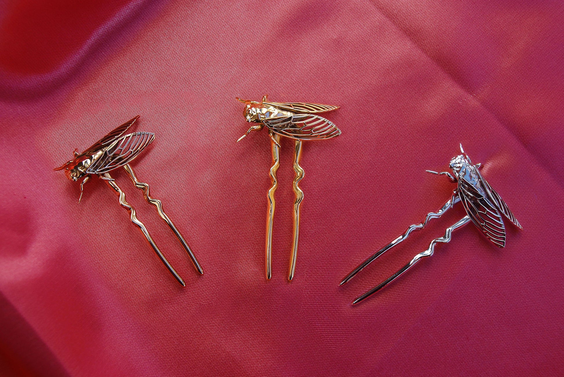 Gold, silver, and bronze cicada hair pins arranged on flowing silk fabric, emphasizing the metallic tones and intricate insect-inspired design against a soft, luxurious backdrop.