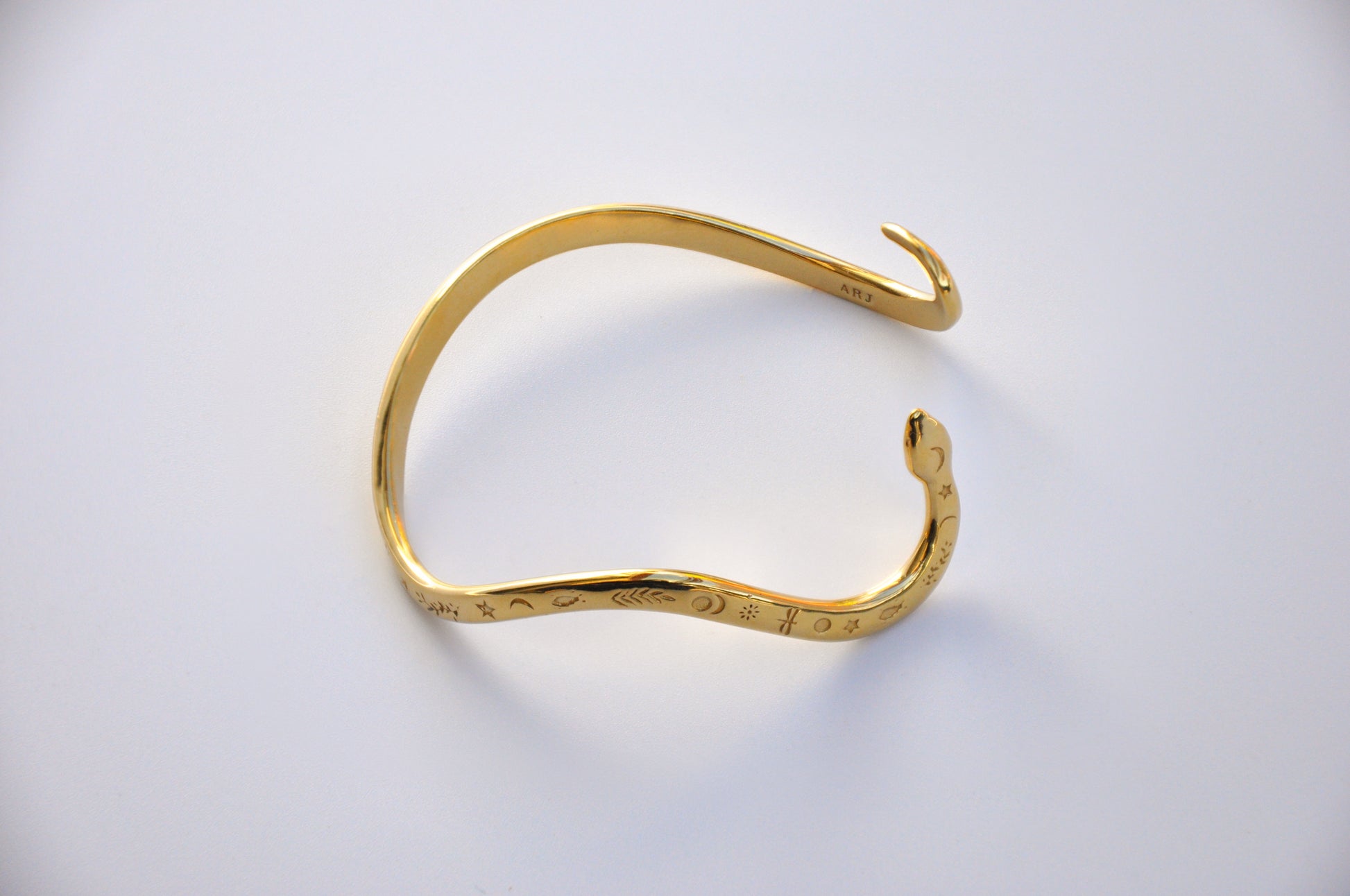Top‑view photograph of a polished gold snake bracelet on a white background, showing engraved celestial details.