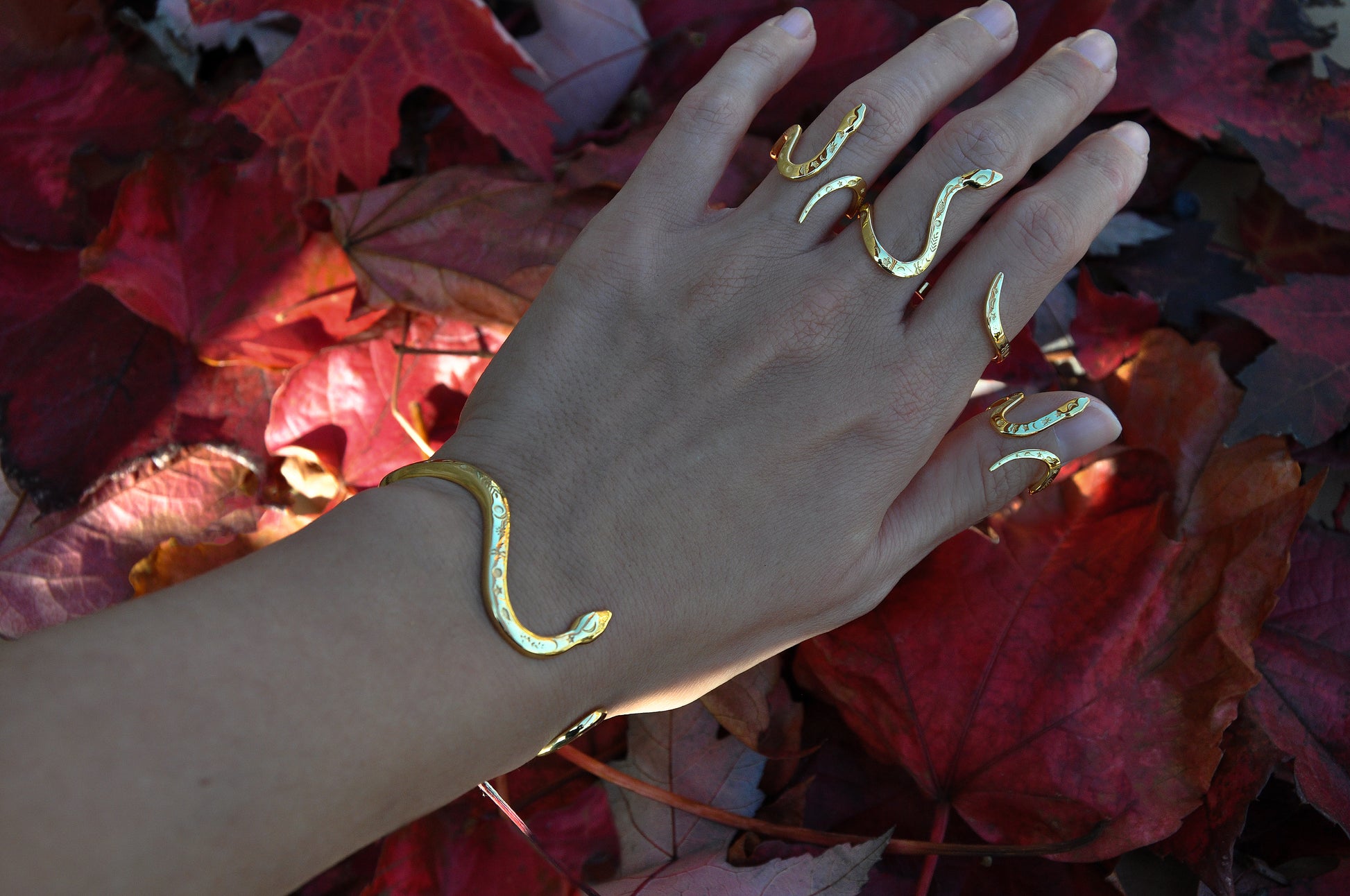 Hand adorned with gold snake rings and bracelet, photographed among red autumn leaves, designed in Italy.