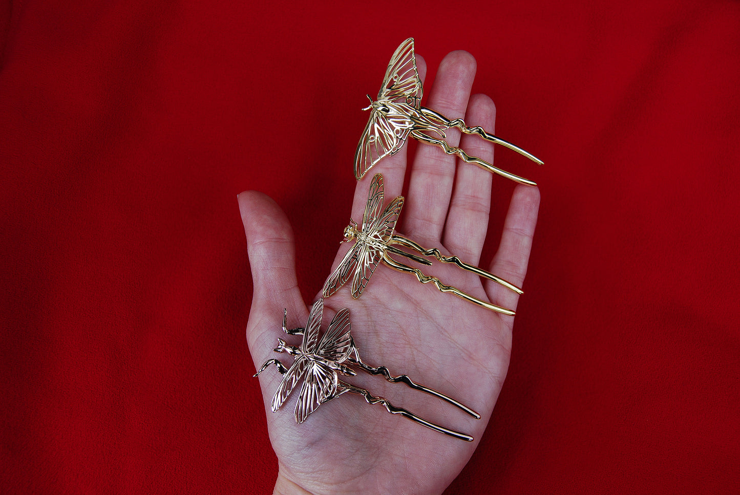 Hand holding three insect-themed hair pins: a 14k gold-plated Luna Moth, a polished bronze Dragonfly, and a 14k rose gold-plated Mantis, showcasing intricate detailing and metallic finishes.