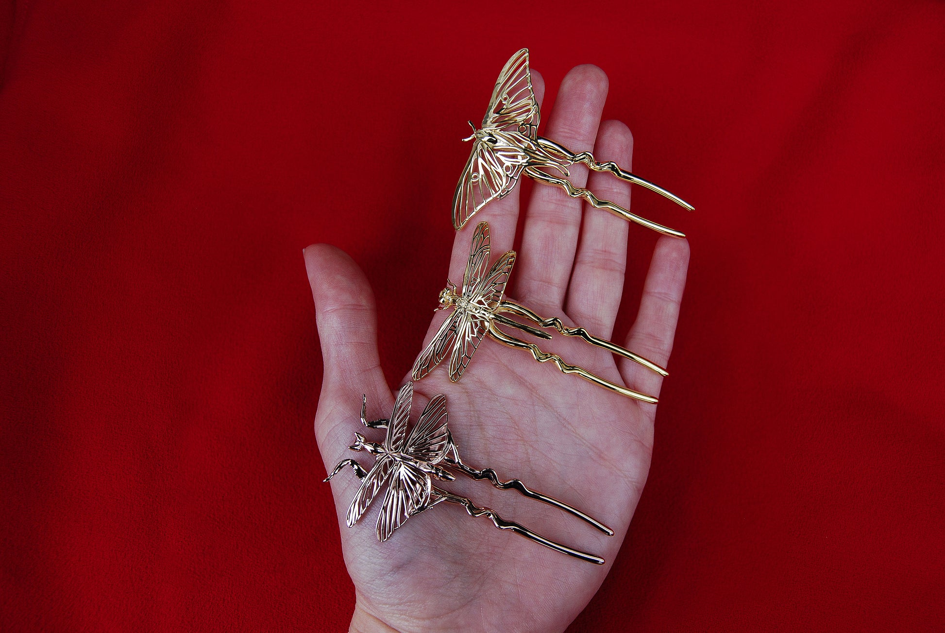 Hand holding three insect-themed hair pins: a 14k gold-plated Luna Moth, a polished bronze Dragonfly, and a 14k rose gold-plated Mantis, showcasing intricate detailing and metallic finishes.