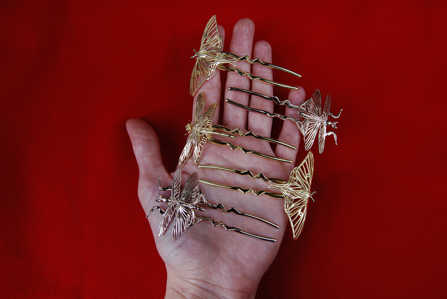 Hand holding three insect-inspired hair pins: one Luna Moth in 14k gold plating, one Luna Moth in solid 14k gold, and one Mantis in 14k rose gold plating, highlighting their intricate craftsmanship and luminous finishes.