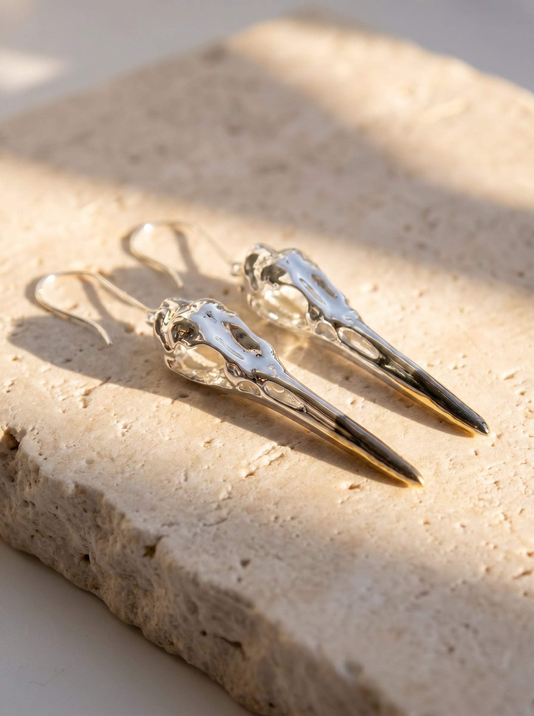 Heron Skull Earrings resting on stone surface, highlighting anatomical engraving and tonal interplay between silver, rhodium, and gold.