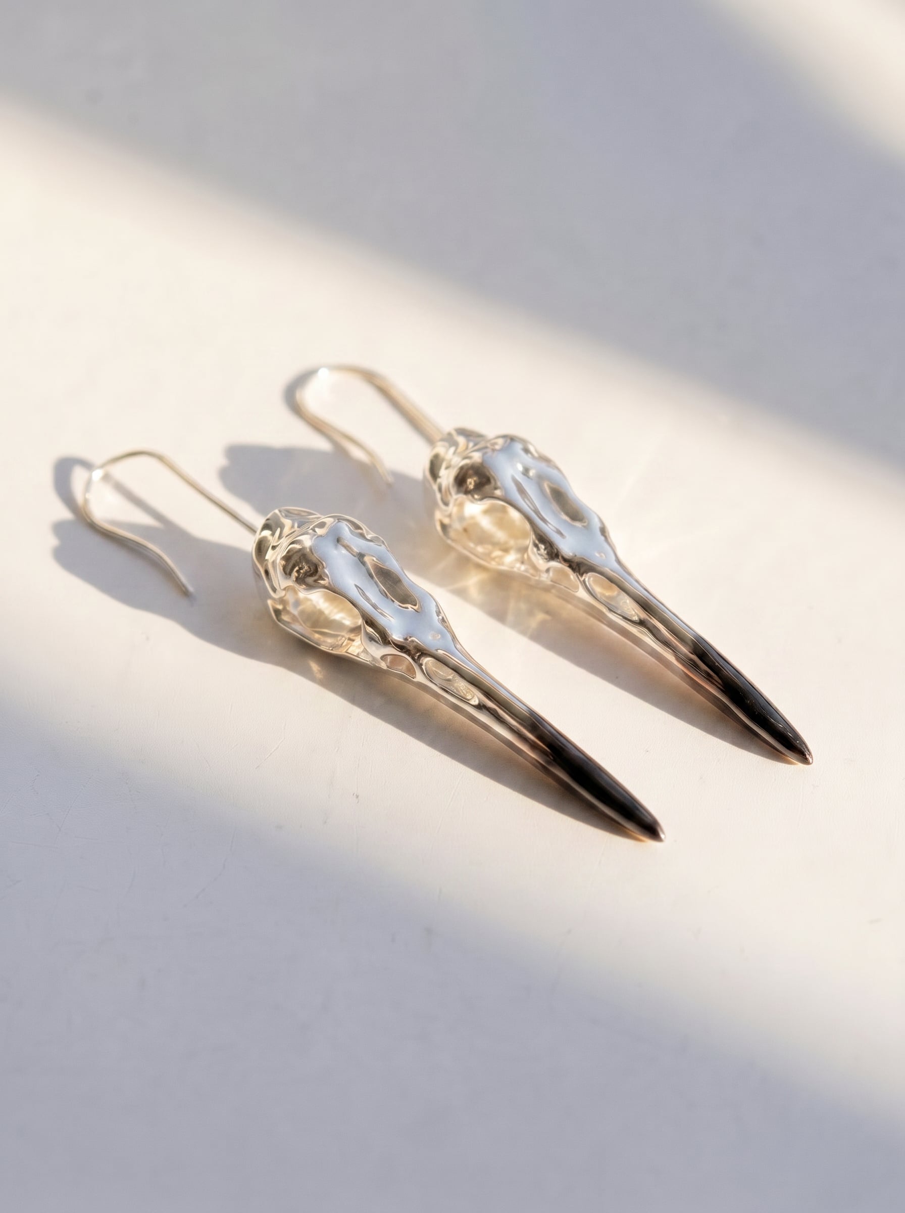 Heron Skull Earrings in sterling silver with black rhodium and gold accents, photographed in sunlight to reveal elongated sculptural form and polished contrast.
