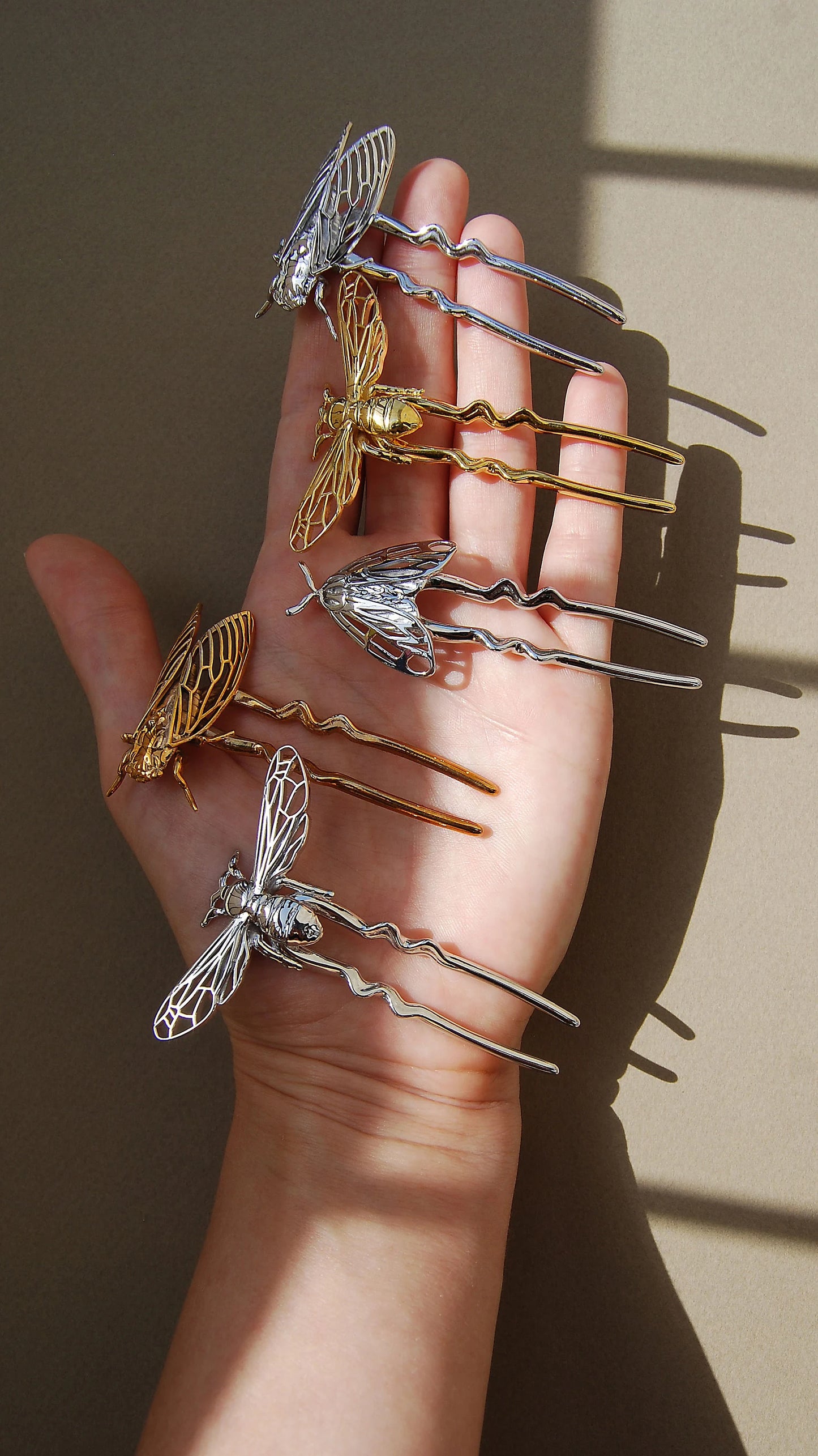 Hand holding a collection of insect-inspired hair pins, including a 14k gold bee pin, silver cicada pin, tiger moth pin, silver bee pin, and silver tiger moth pin, showcasing detailed craftsmanship and nature-themed jewelry design.