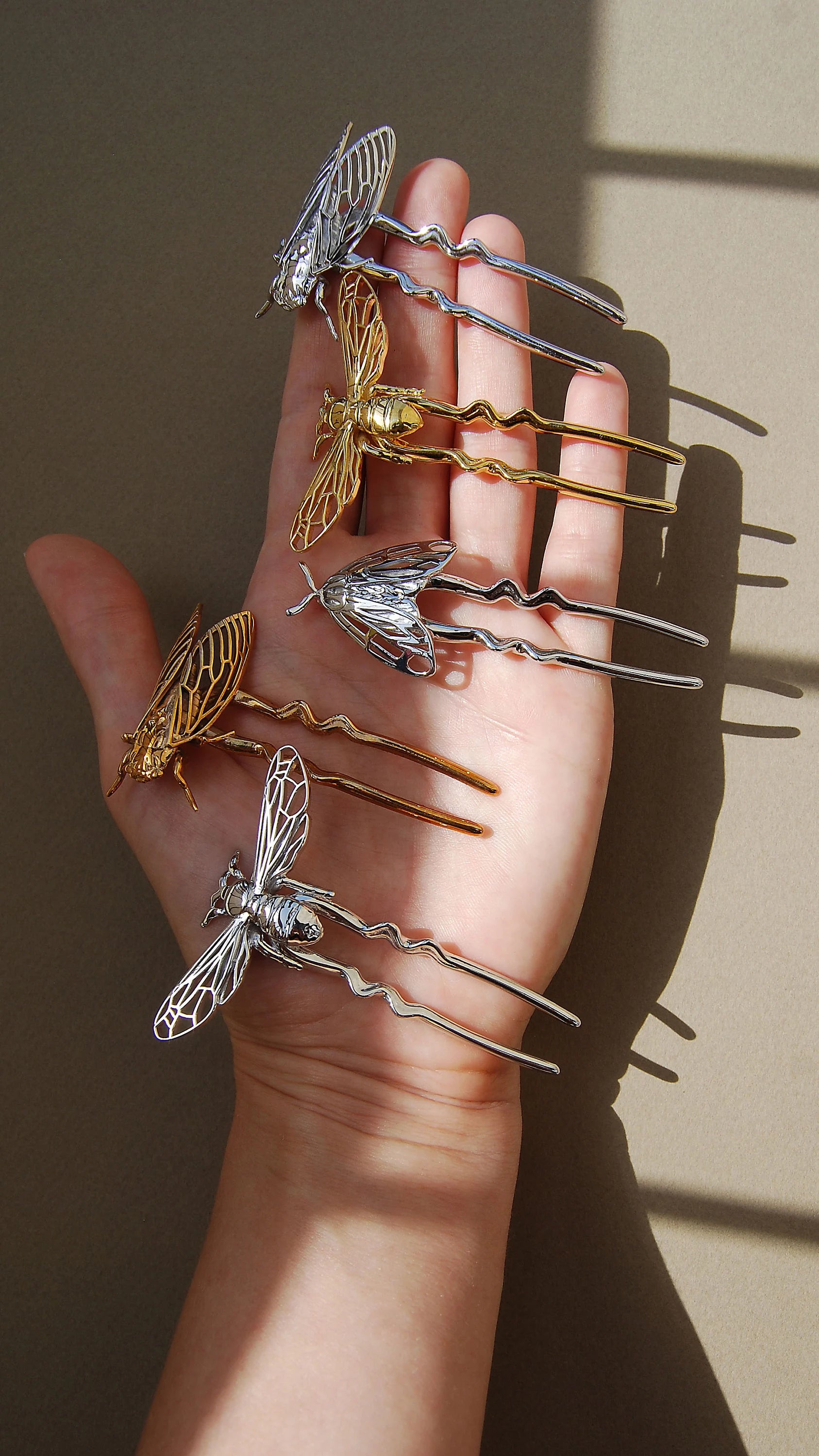 Hand holding a collection of insect-inspired hair pins, including a 14k gold bee pin, silver cicada pin, tiger moth pin, silver bee pin, and silver tiger moth pin, showcasing detailed craftsmanship and nature-themed jewelry design.