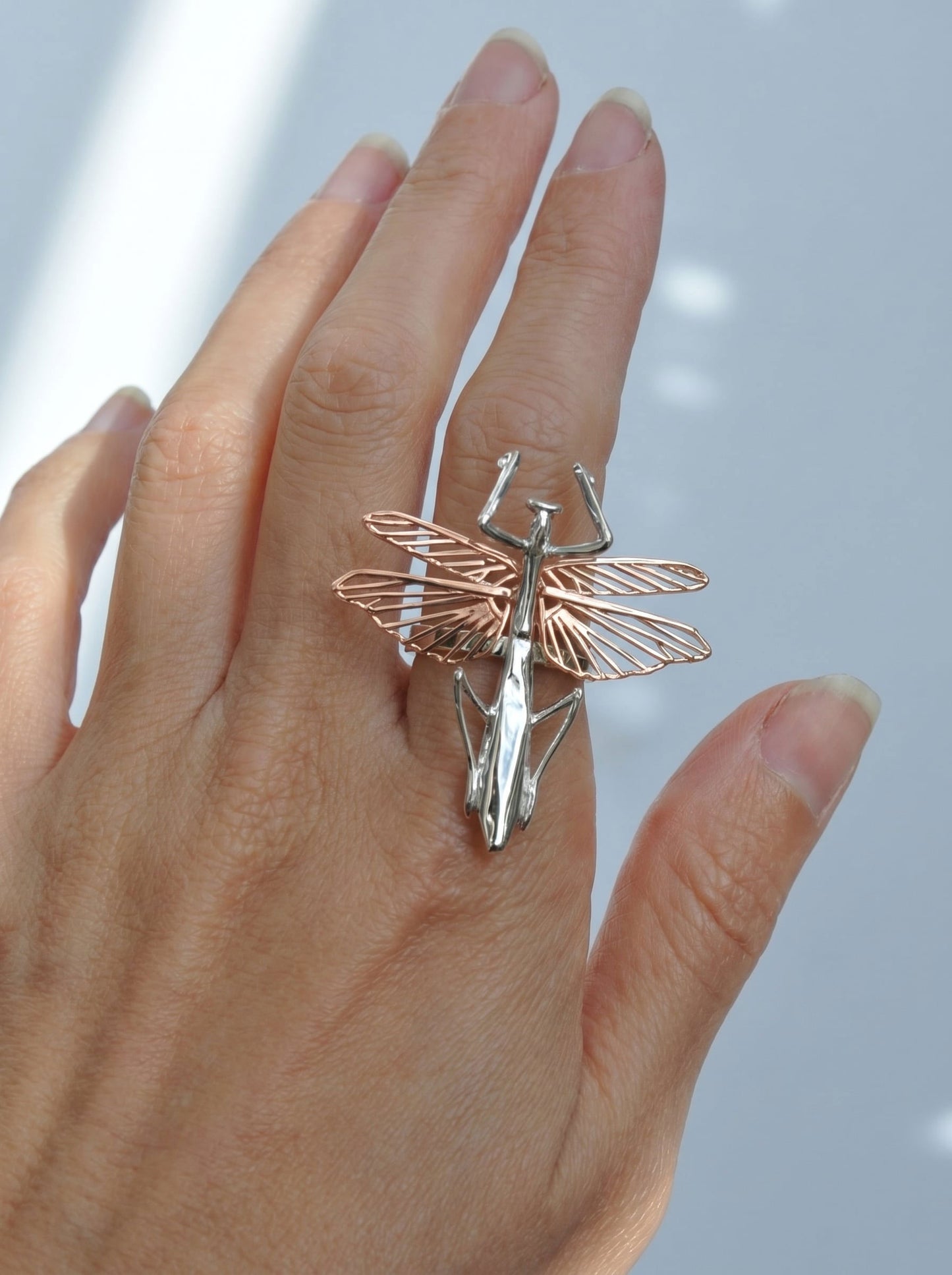 Praying mantis ring with a sterling‑silver body and rose‑gold wings shown on the hand.
