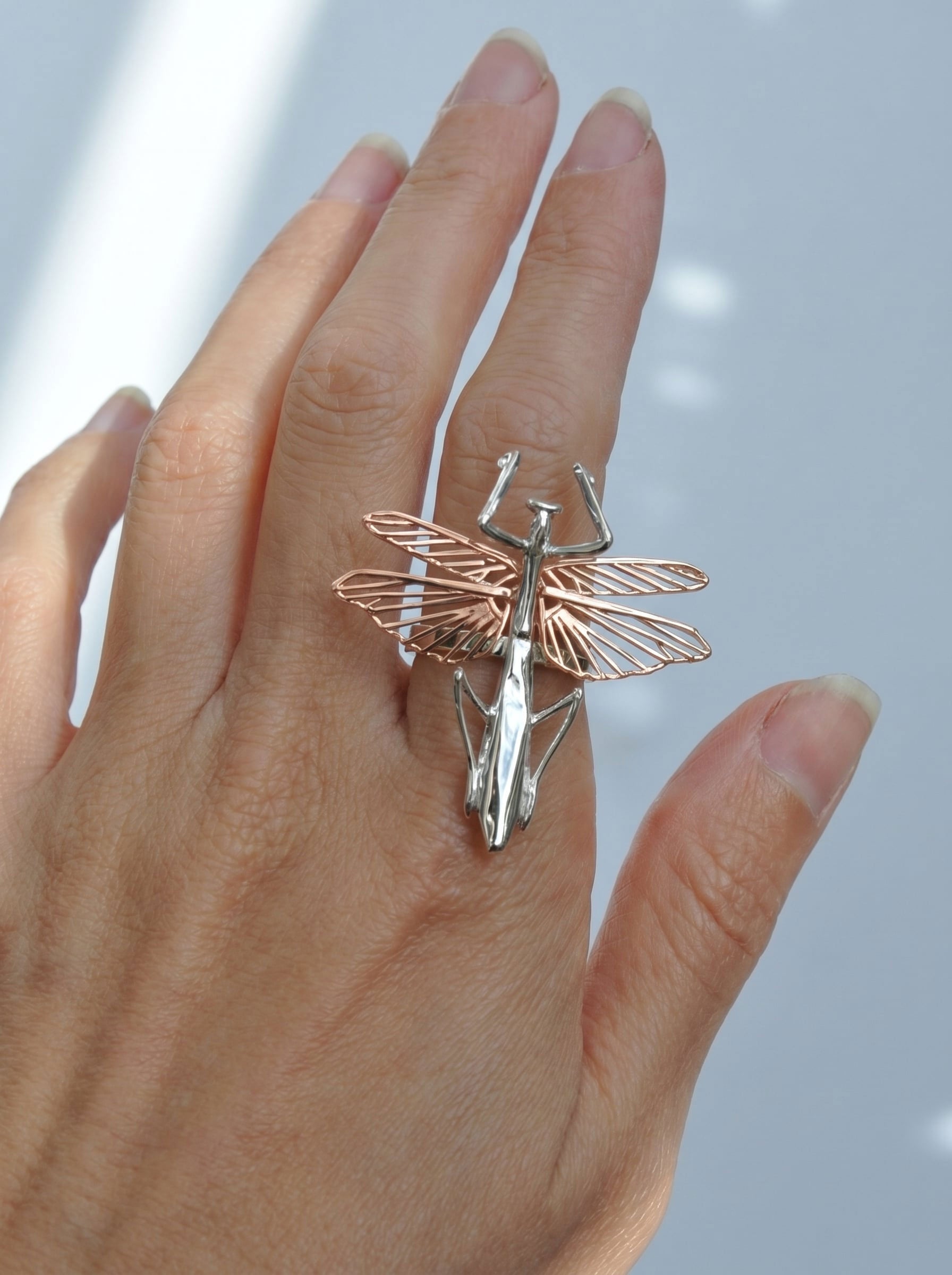 Praying mantis ring with a sterling‑silver body and rose‑gold wings shown on the hand.