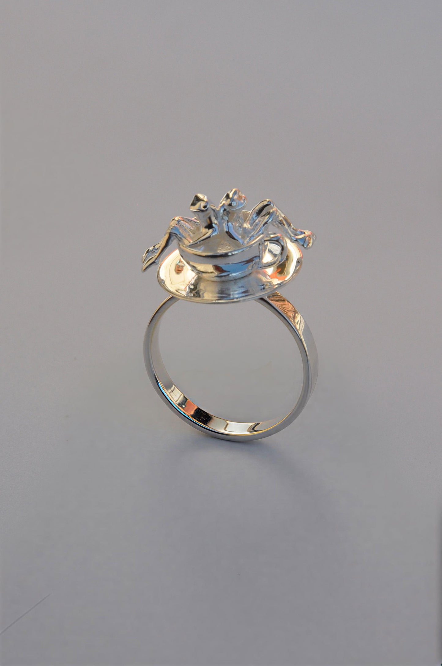 Miniature carousel‑style ring with two detailed frog figures inside a silver teacup set on a spinning saucer.