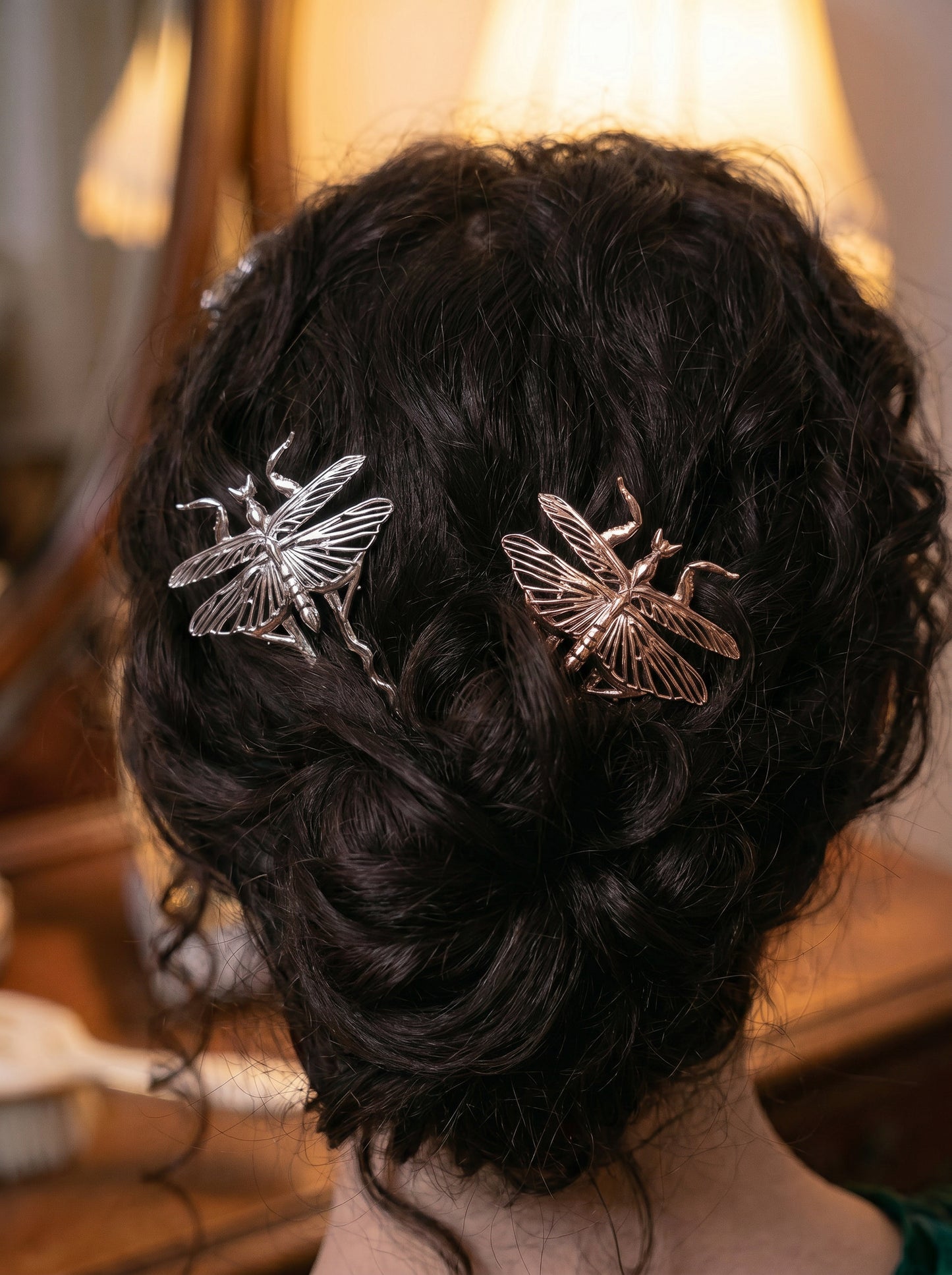 Silver and rose gold Orchid Mantis hairpins styled in dark curly hair under warm vintage lamplight, delicate and surreal.