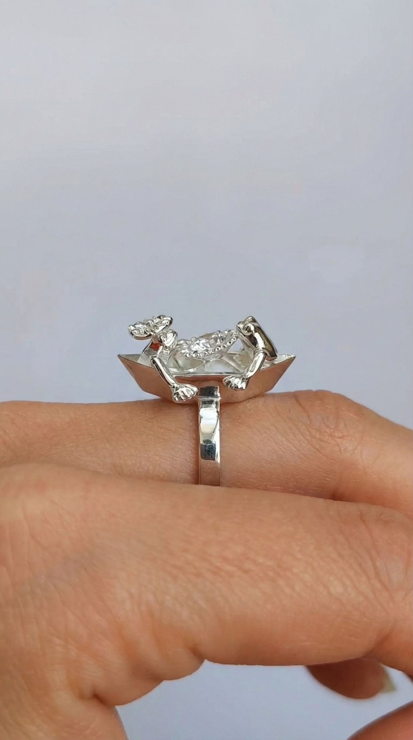 The sterling silver Origami Boat Proposal Ring worn on a finger, showing two tiny frogs and a central clear crystal on the boat‑shaped top.