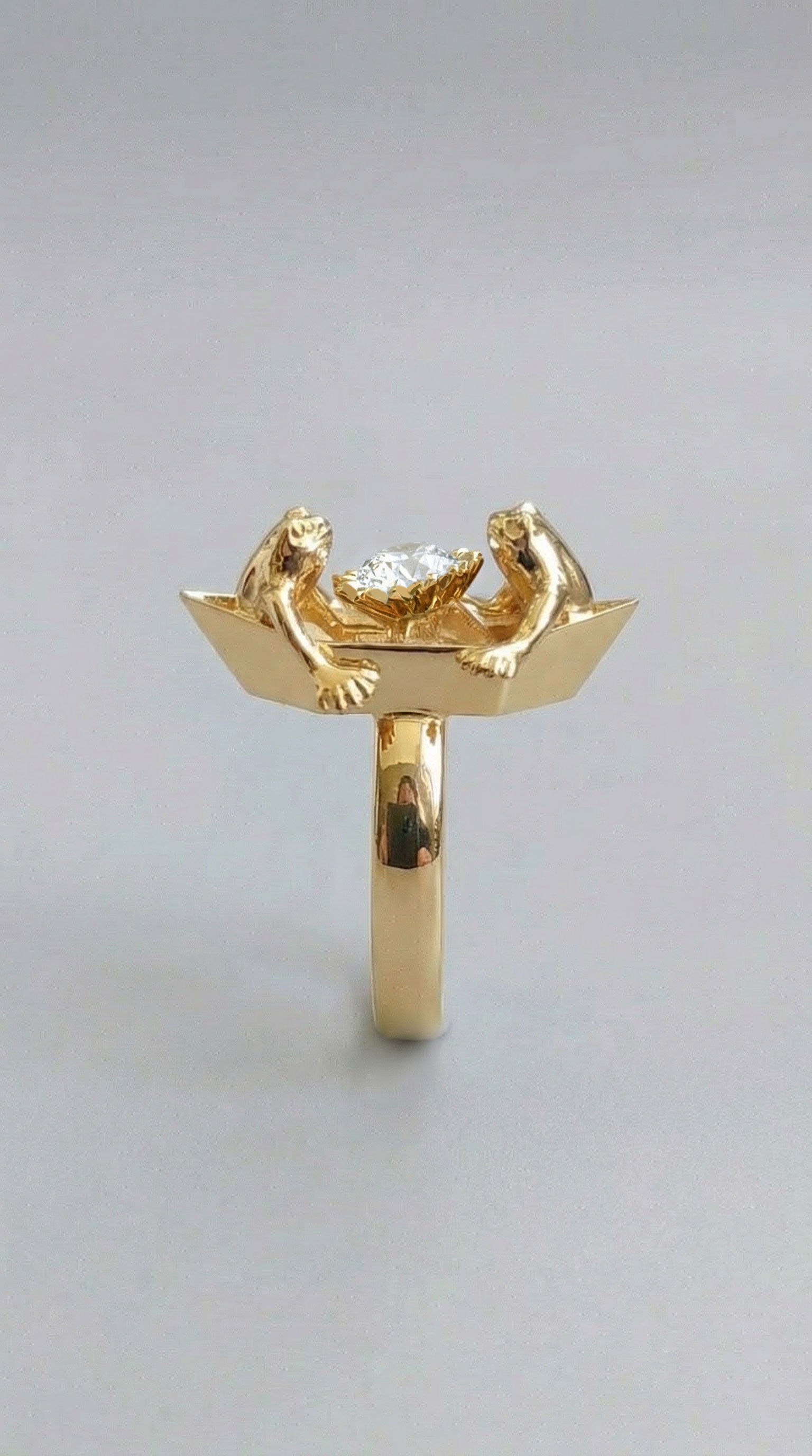 A handcrafted ring featuring two tiny frogs on an origami‑style boat, holding a clear faceted crystal between them, shown in a polished gold finish.