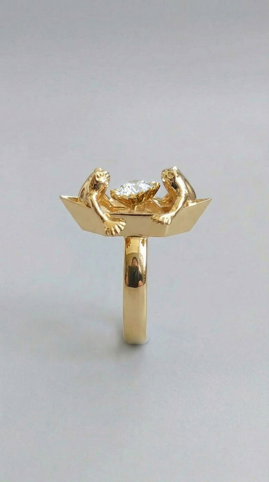 A handcrafted ring featuring two tiny frogs on an origami‑style boat, holding a clear faceted crystal between them, shown in a polished gold finish.