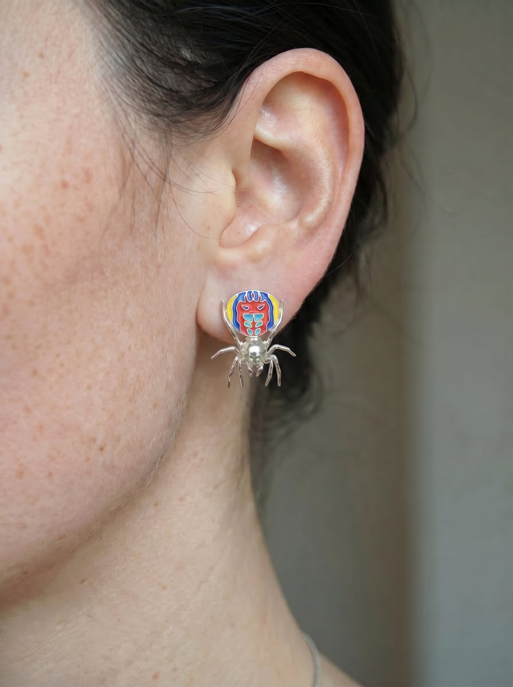Small sterling‑silver peacock spider earring with vibrant enamel detailing worn on the ear.