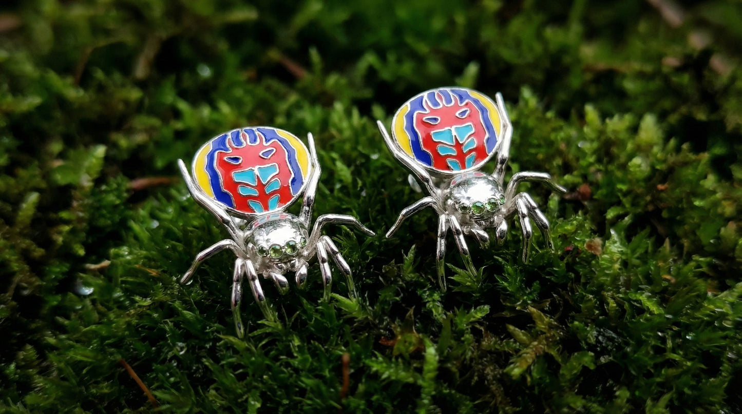 Pair of peacock spider earrings with vibrant enamel detailing displayed on green moss.