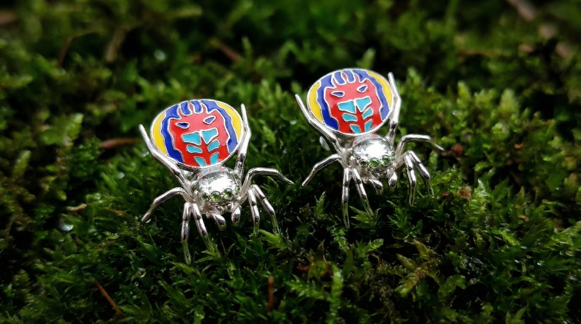 Pair of peacock spider earrings with vibrant enamel detailing displayed on green moss.