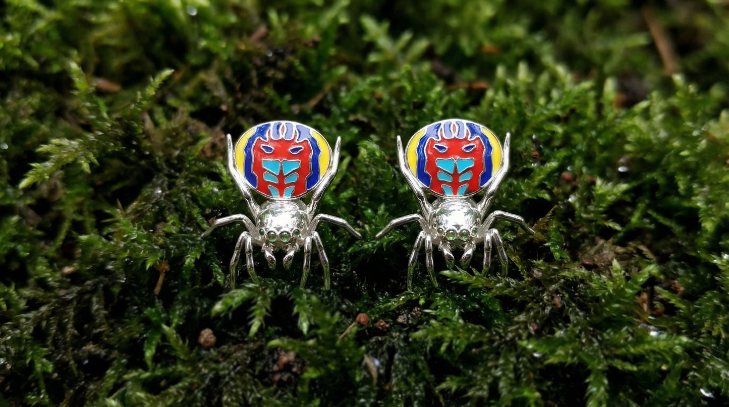 Pair of peacock spider earrings with vibrant enamel detailing displayed on green moss.