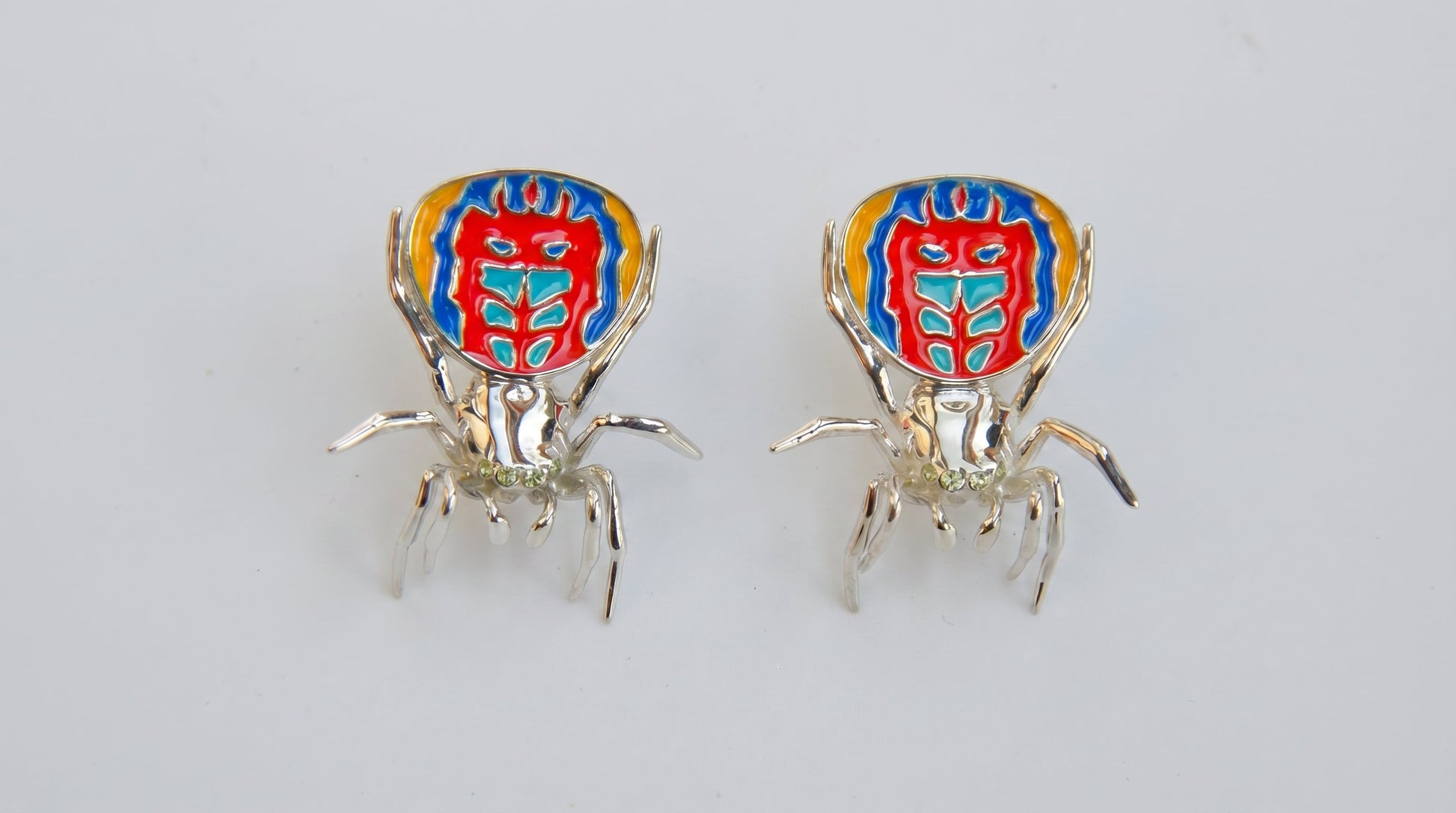 Pair of peacock spider earrings with vibrant enamel detailing displayed on a white background.