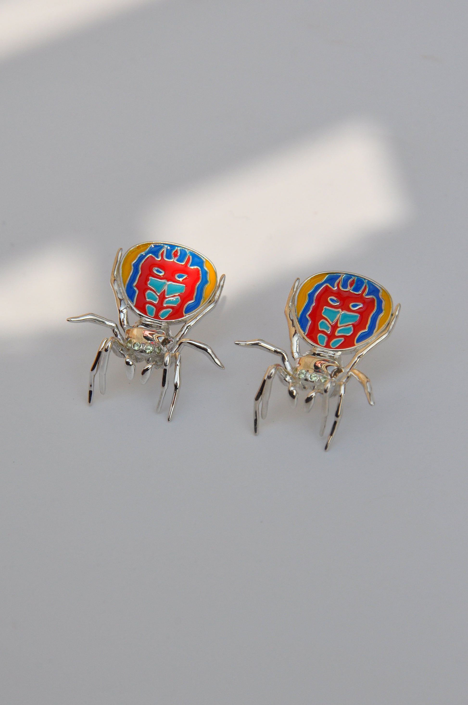 Pair of peacock spider earrings with vibrant enamel detailing displayed on a white background.