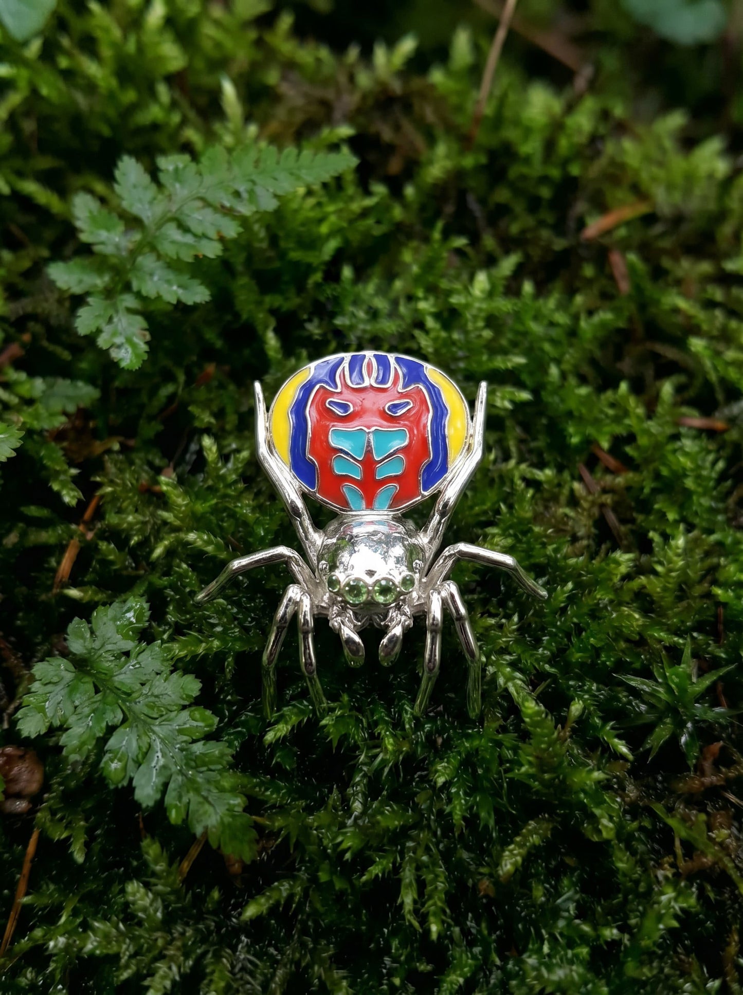 Peacock spider pendant with vibrant enamel abdomen and polished metal legs displayed on green moss.