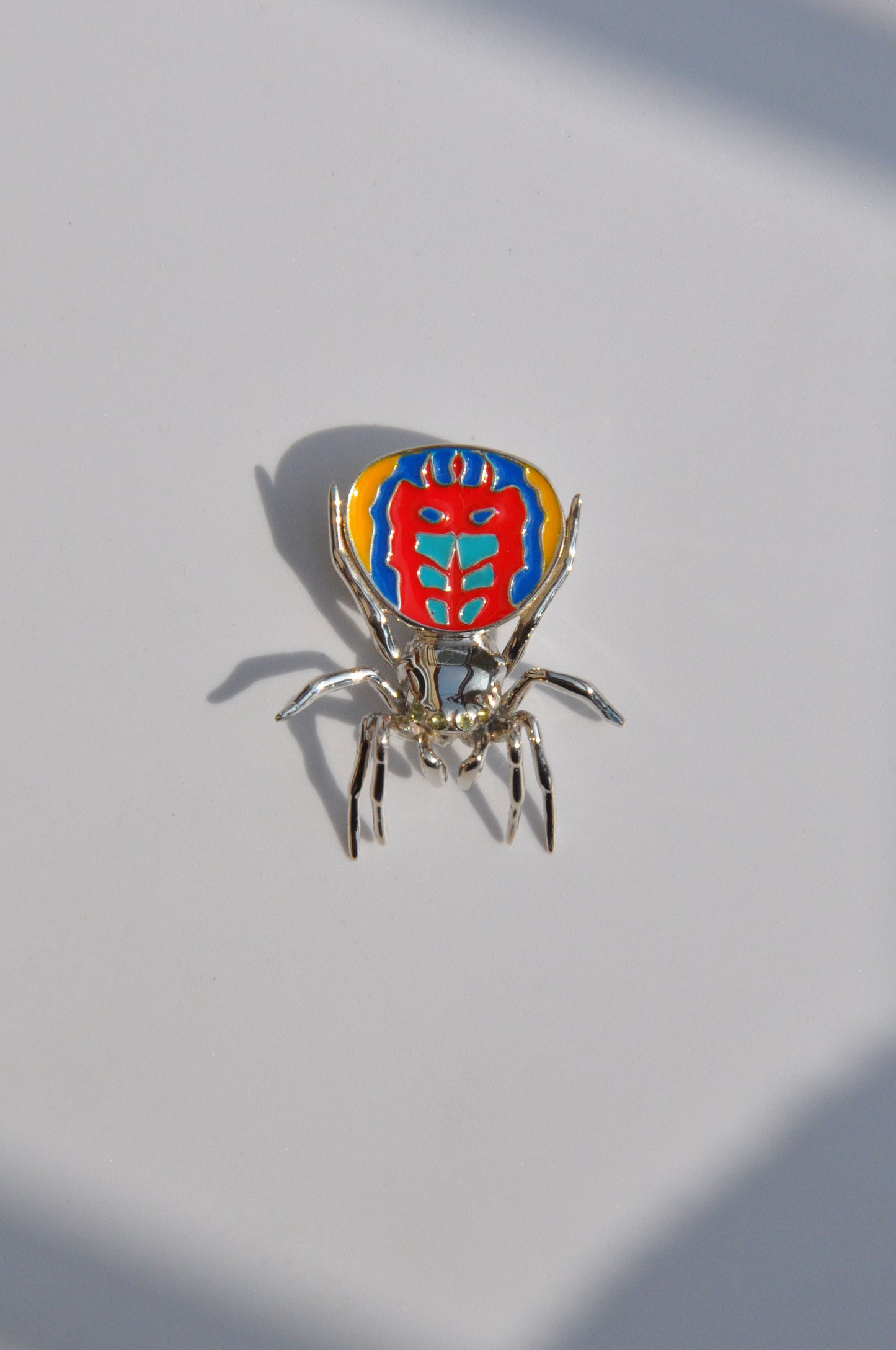 Peacock spider pendant with a polished silverbody and vibrant enamel abdomen displayed on a light surface.