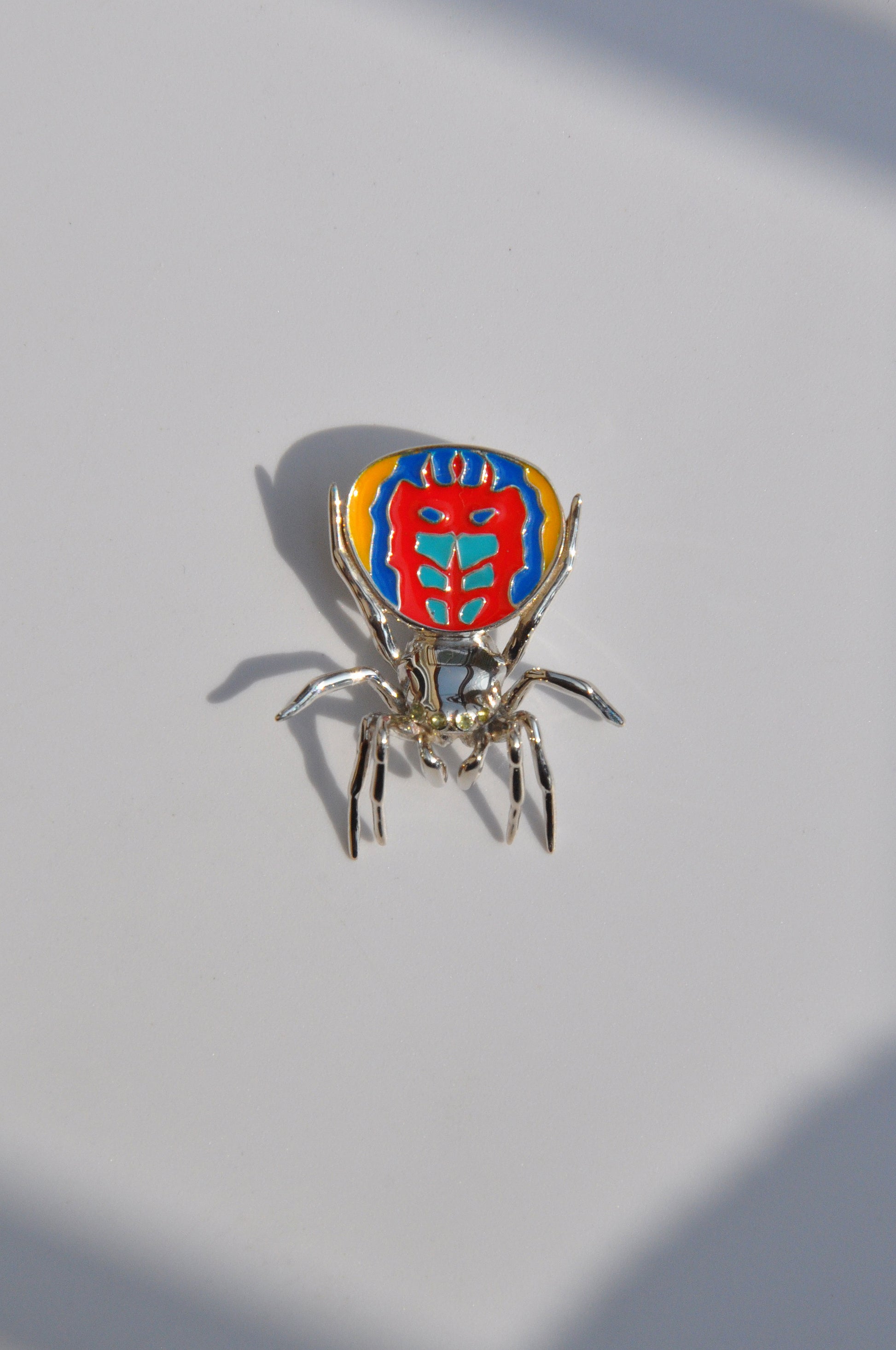 Peacock spider pendant with a polished silverbody and vibrant enamel abdomen displayed on a light surface.