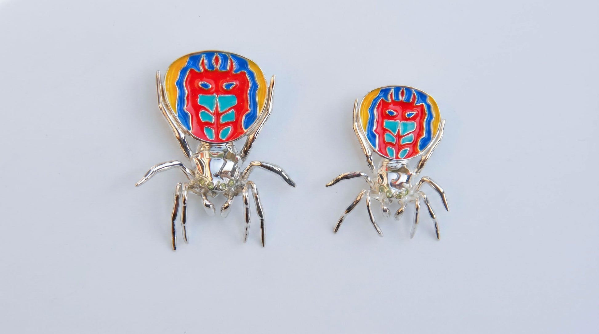 Large and small peacock spider pendants shown side by side with polished metal bodies and vibrant enamel patterns.