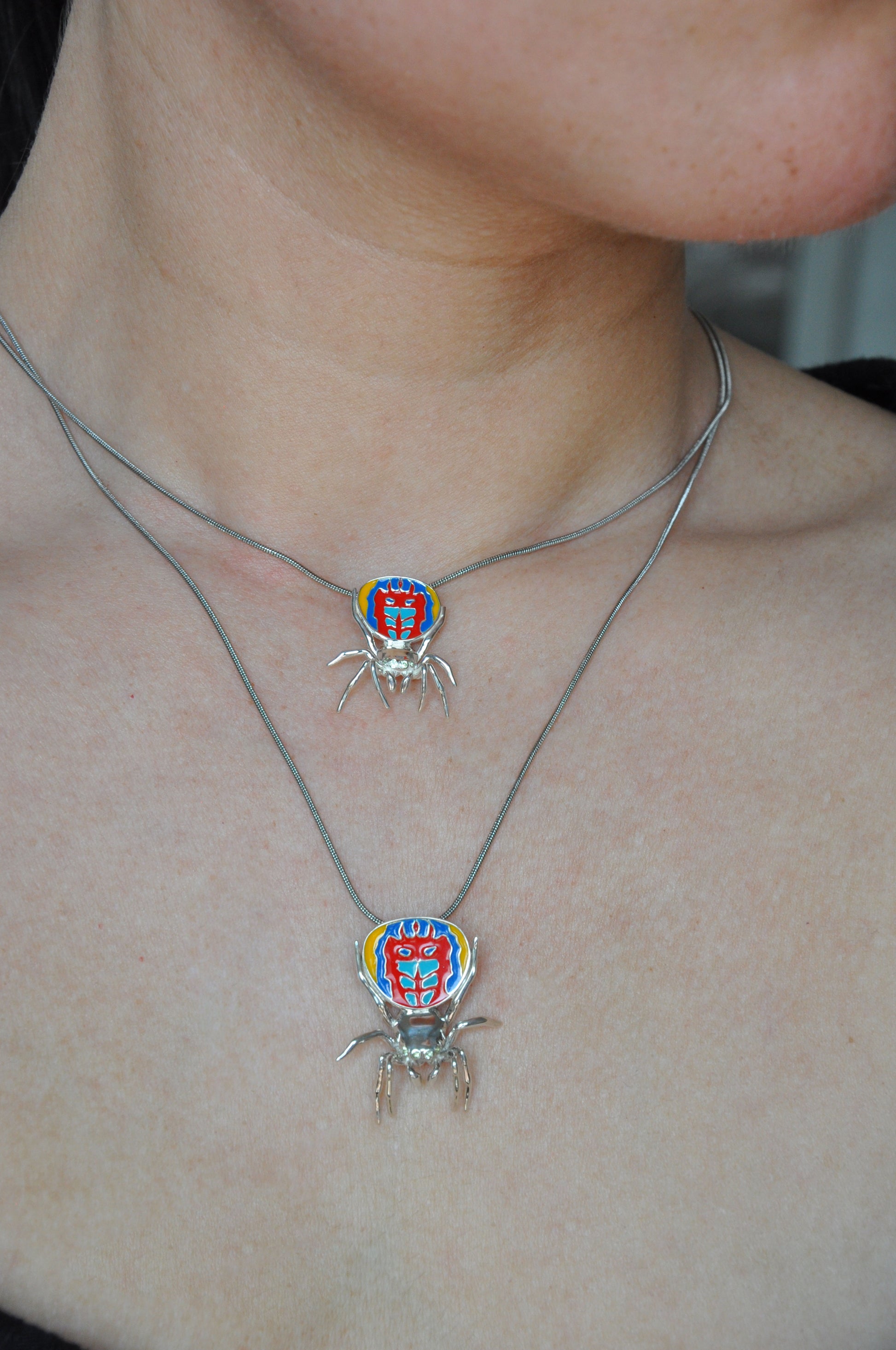 Two sterling‑silver peacock spider pendants in small and large sizes worn on layered chains, featuring vibrant enamel detailing.