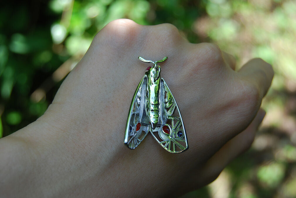 Back view of Picasso Moth pendant resting on hand with green forest background, showing sterling silver craftsmanship and rhodium finish