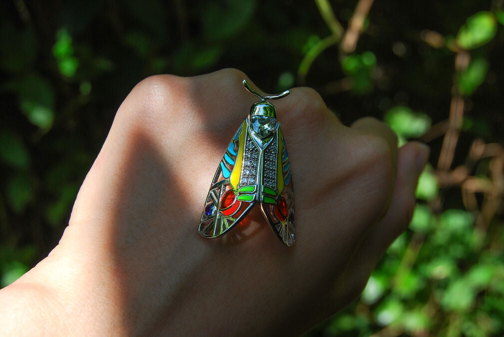 Picasso Moth pendant with colorful enamel and zirconia stones resting on hand against green forest background