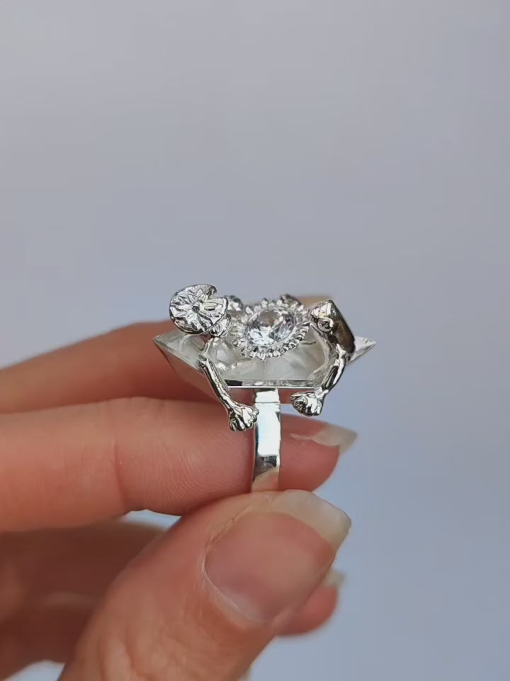Video showing a sterling silver ring worn on a finger, with two sculpted frogs on a boat‑shaped top holding a clear faceted crystal.